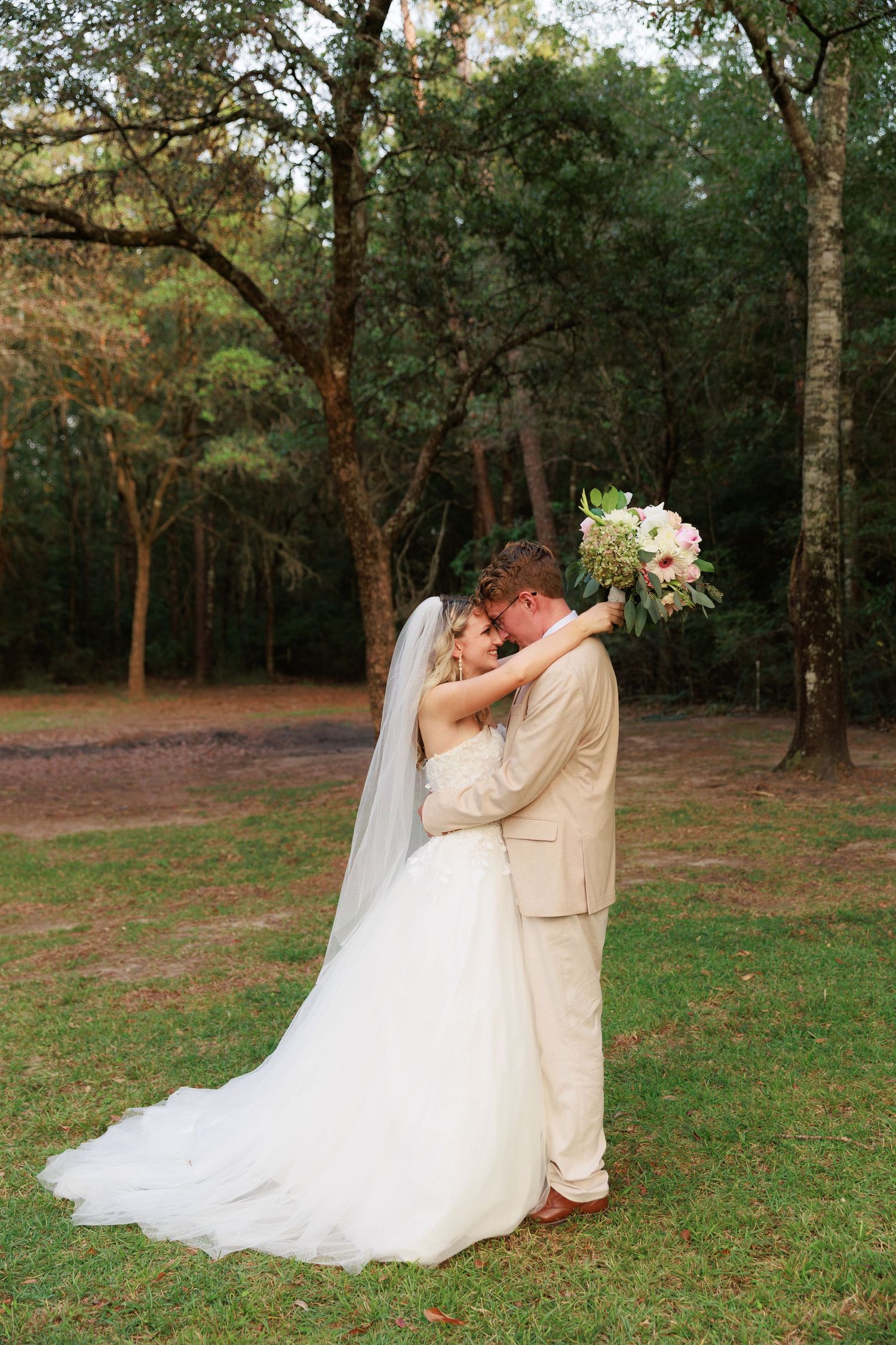 A couple in wedding attire shares romantic moments in a wooded outdoor setting with tall trees and grass.
