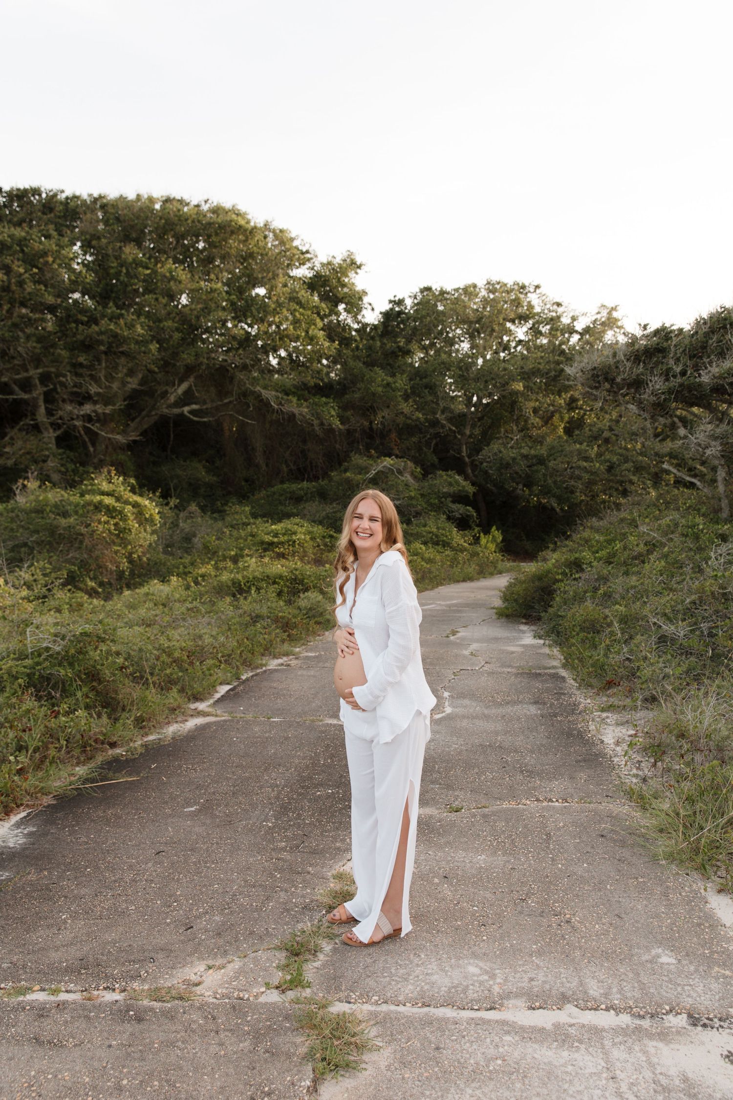 A person in a flowing white dress poses gracefully on a winding dirt path surrounded by green foliage.