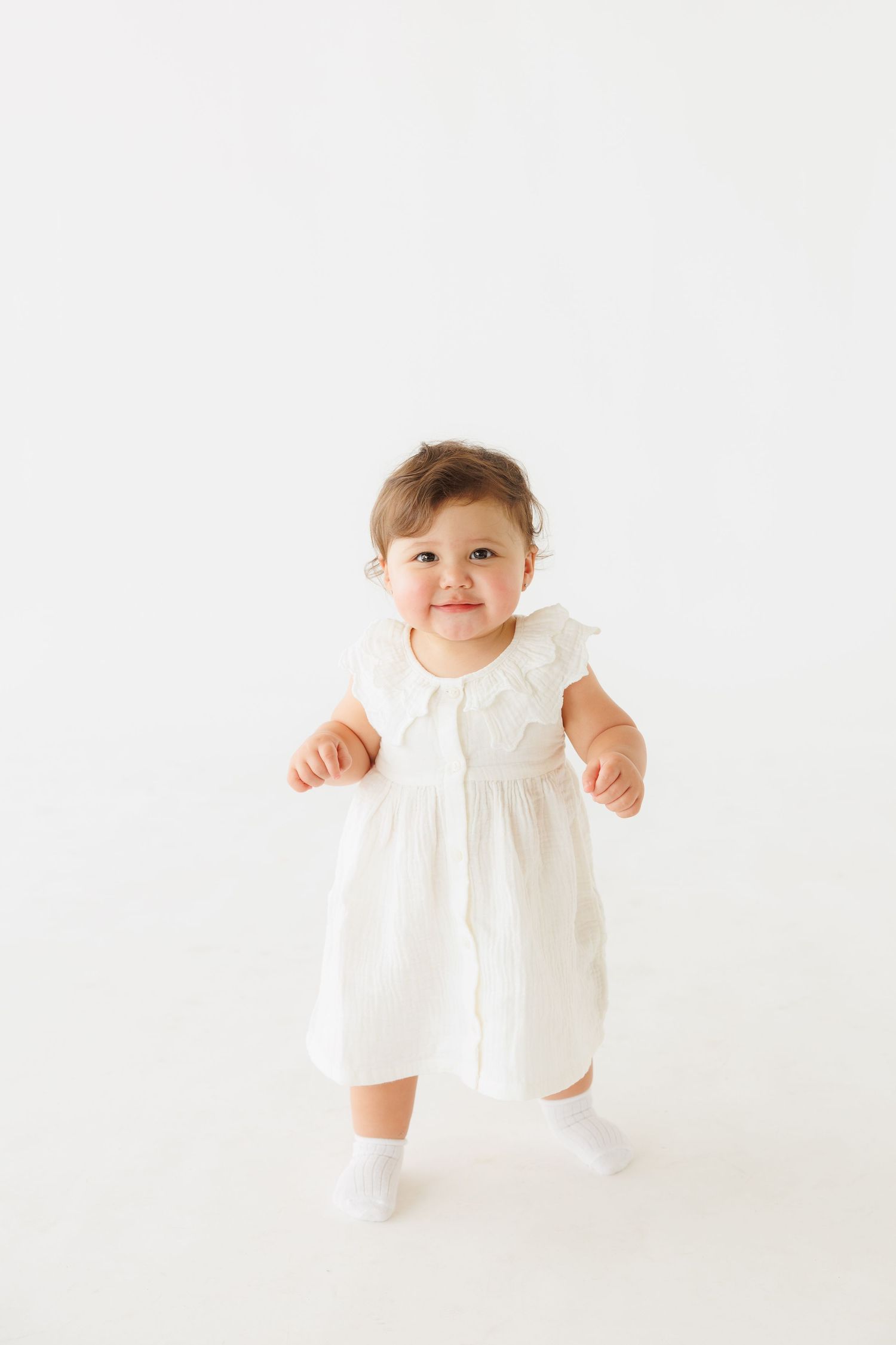Smiling baby girl in white dress and socks standing on white background.