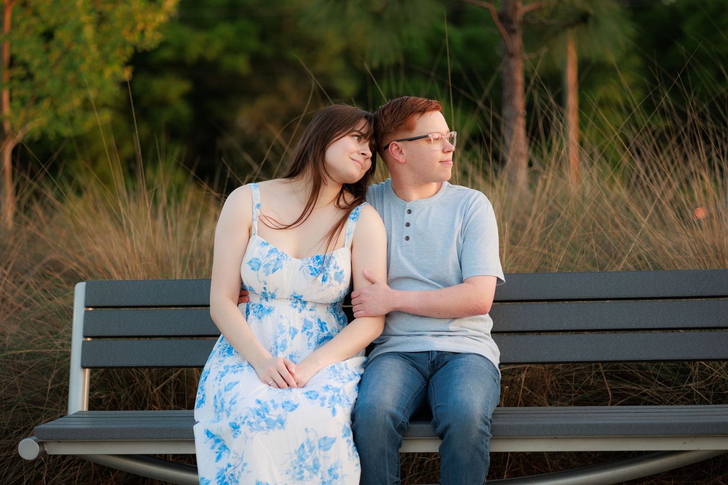 Couple sitting on a park bench at golden hour, woman in floral dress leaning on man in blue shirt.