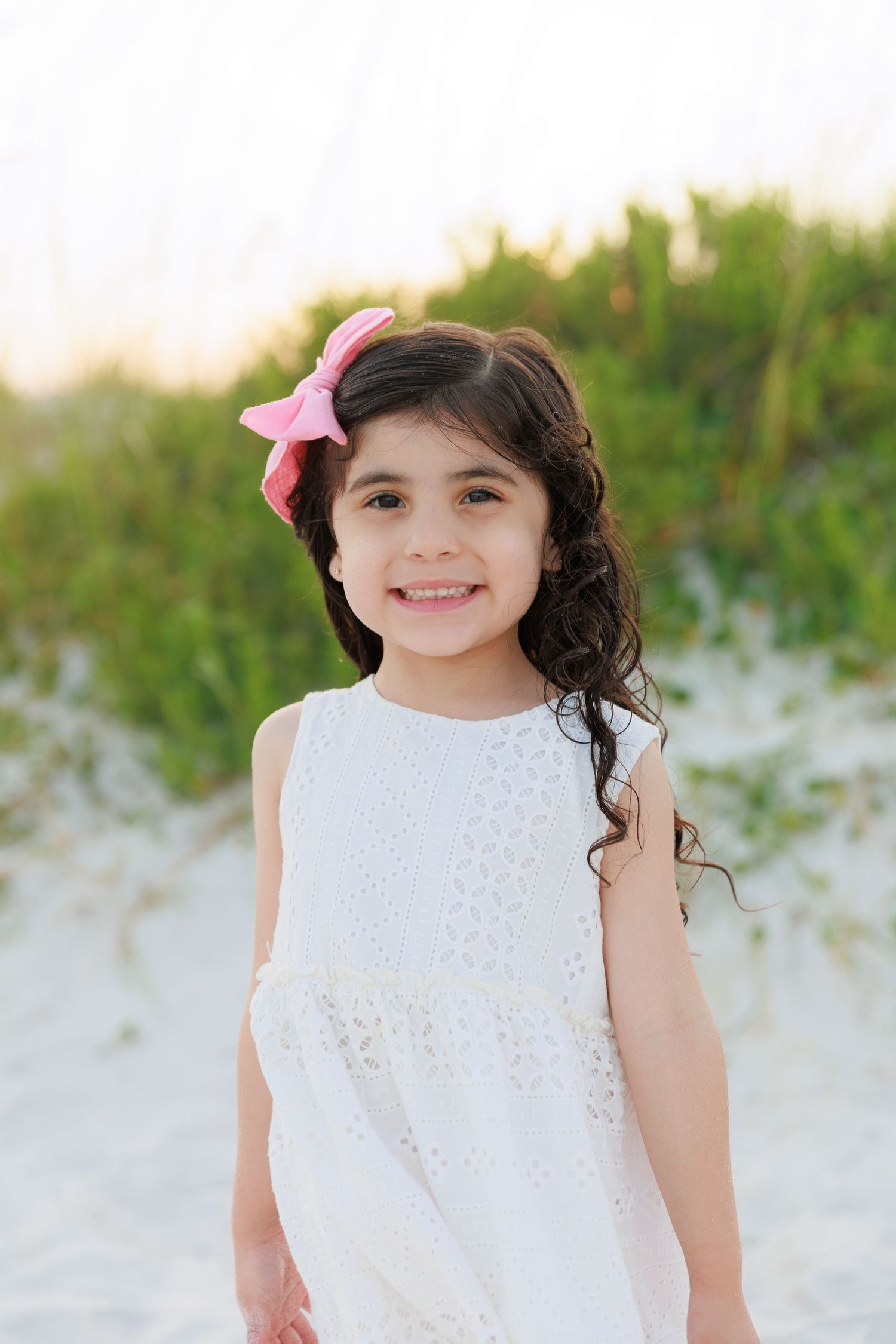 A smiling young girl in a white eyelet dress and pink bow poses on a sandy beach with green dunes behind her.
