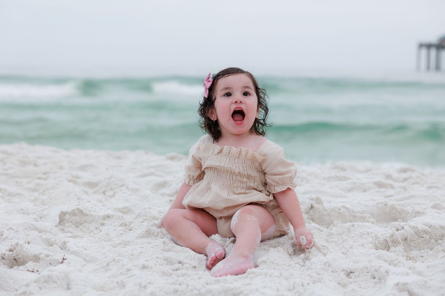 Toddler girl with curly hair sitting on white sand beach, mouth open in excitement, wearing a beige smocked outfit.