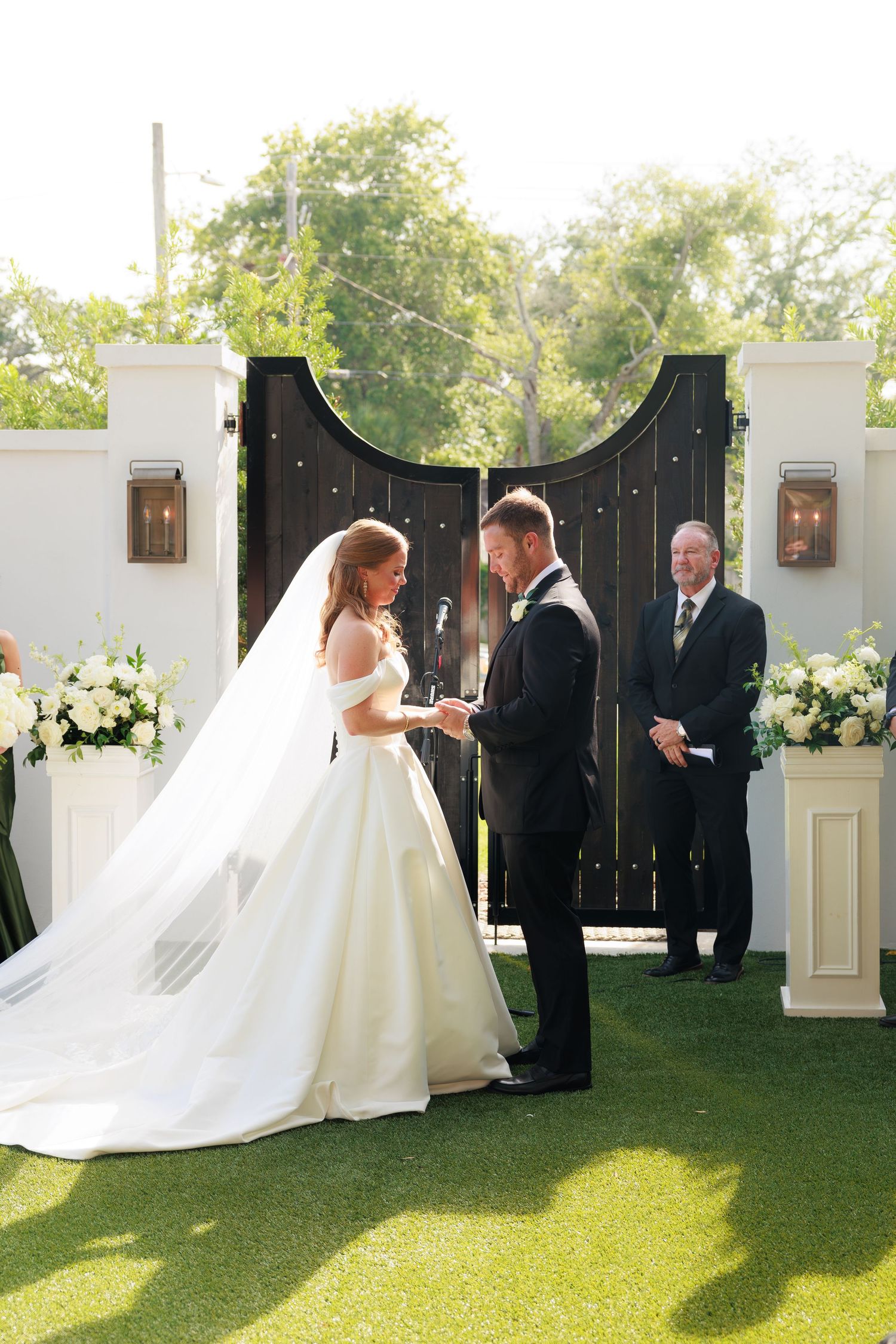 Bride and groom exchange rings outdoors at elegant wedding ceremony with white floral arrangements and dark wooden gates.