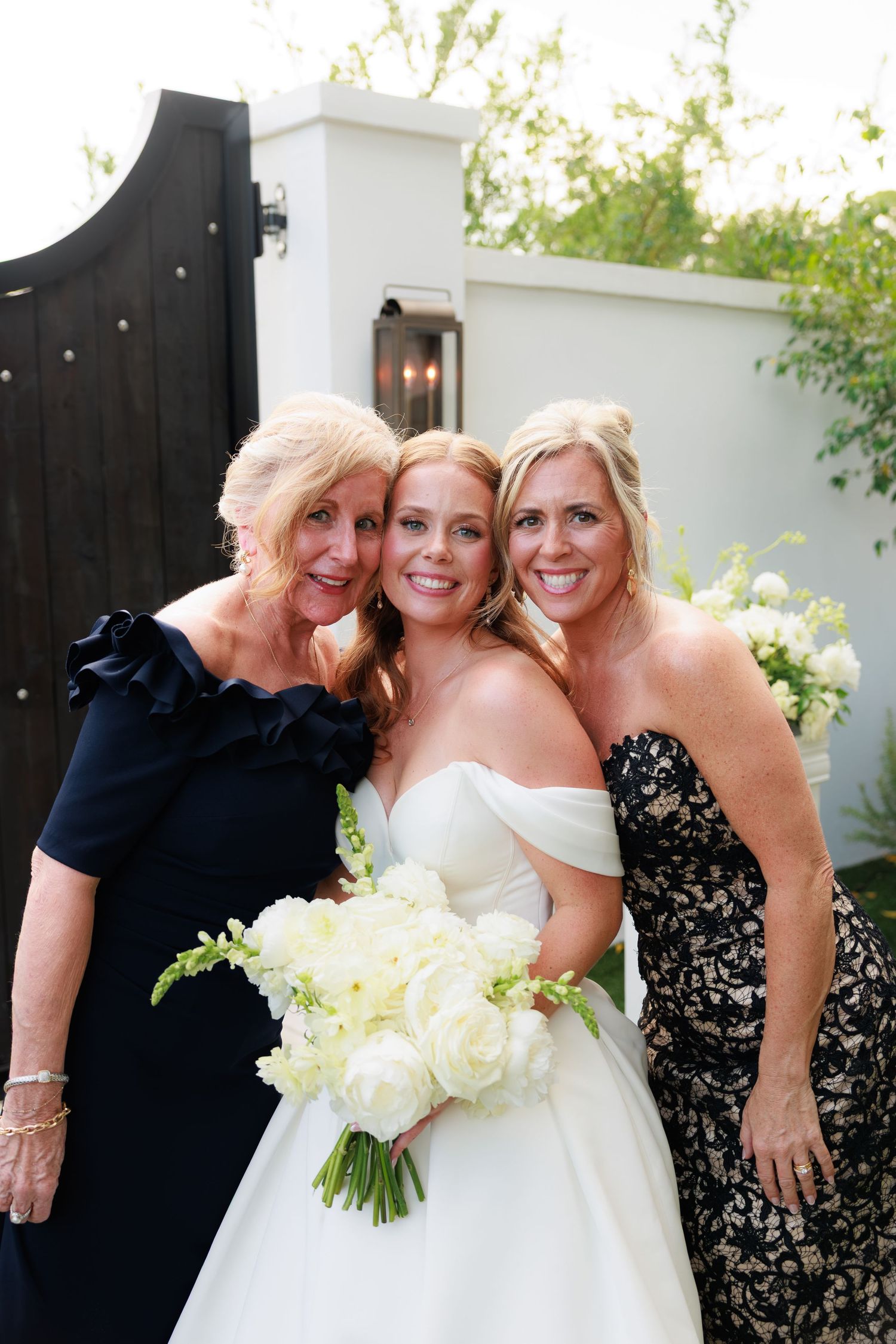 Bride in white gown holds bouquet with two blonde women in black dresses smiling outdoors at wedding celebration.