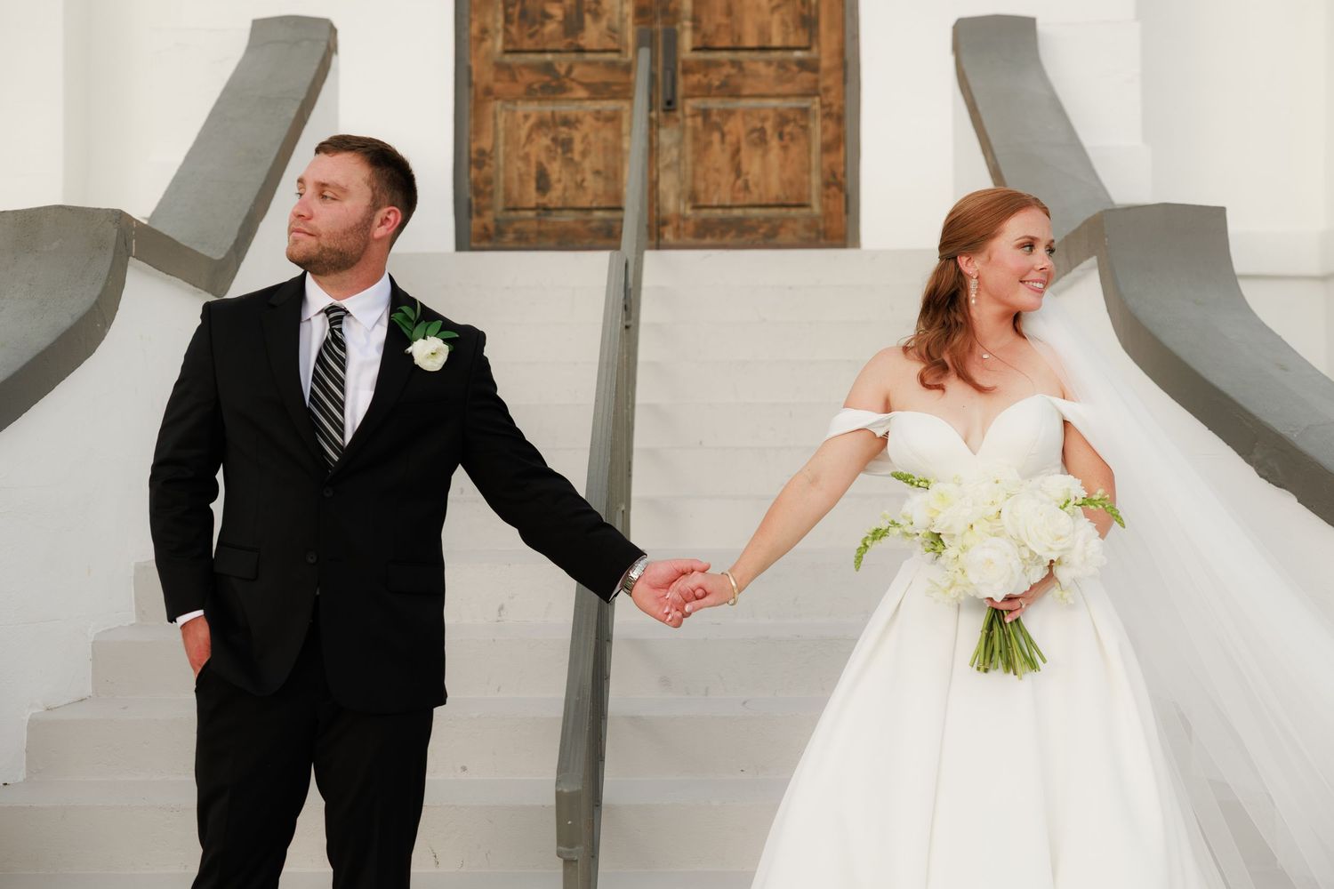 Bride in white ballgown and groom in black suit holding hands at elegant white staircase.