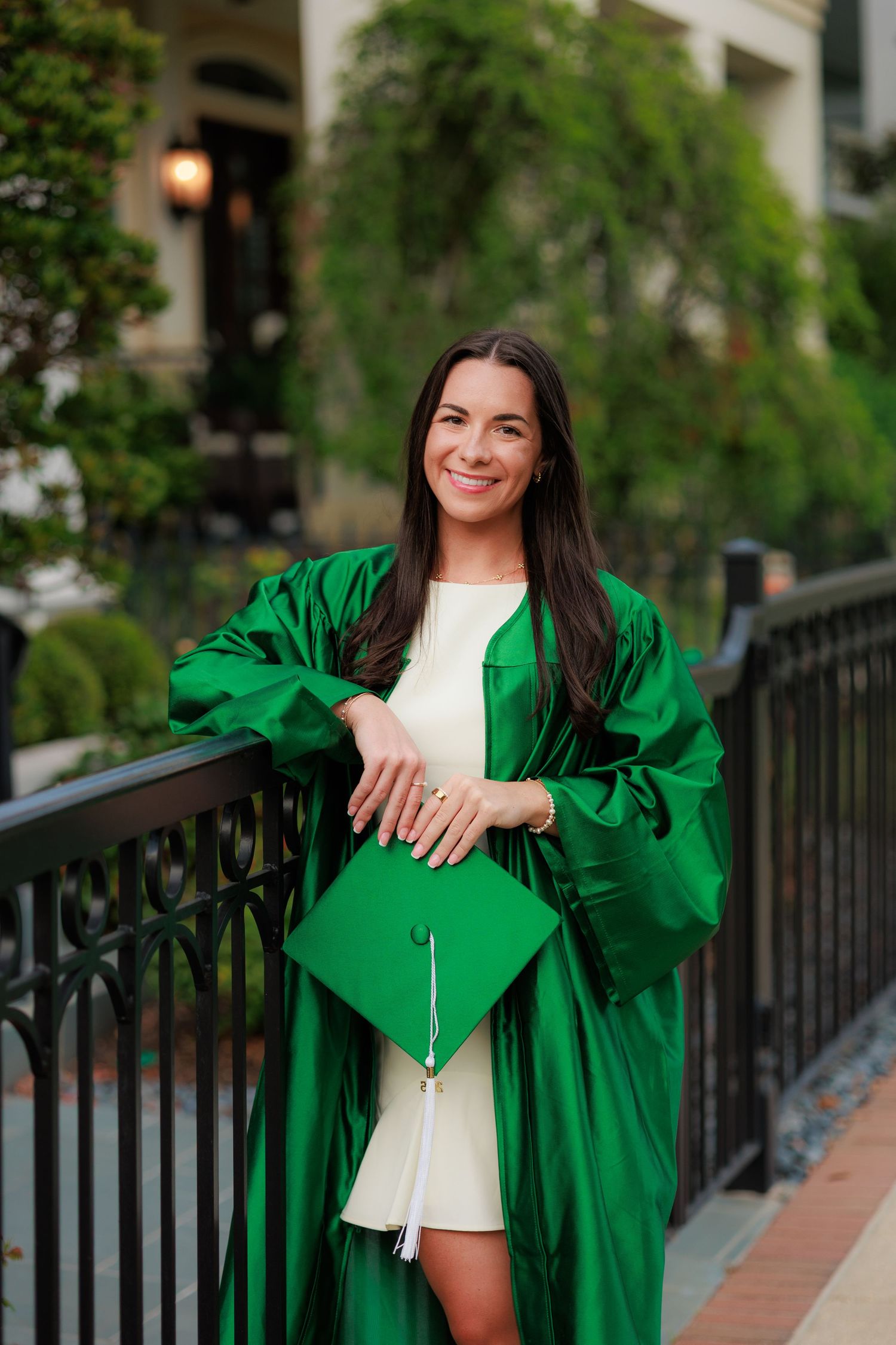 Young woman smiling in green graduation gown and cap, posing by iron railing outdoors.