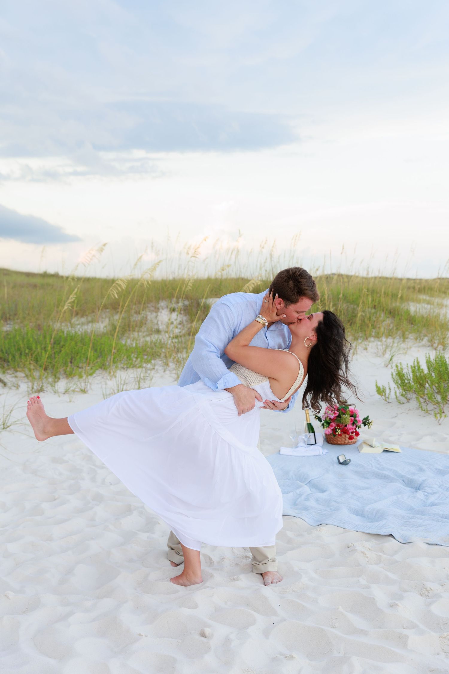 A romantic beach scene with a couple in blue and white sharing an intimate moment on a white blanket.