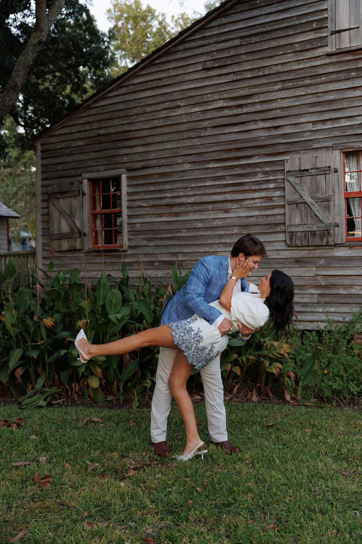 A couple shares a romantic dip pose in front of a rustic wooden barn with red windows at dusk.
