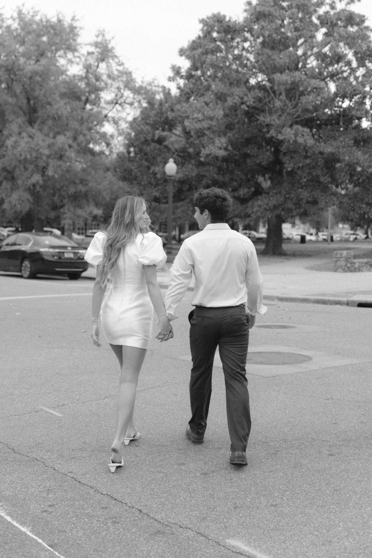 A black and white photo of a couple walking hand in hand through a tree-lined parking area.