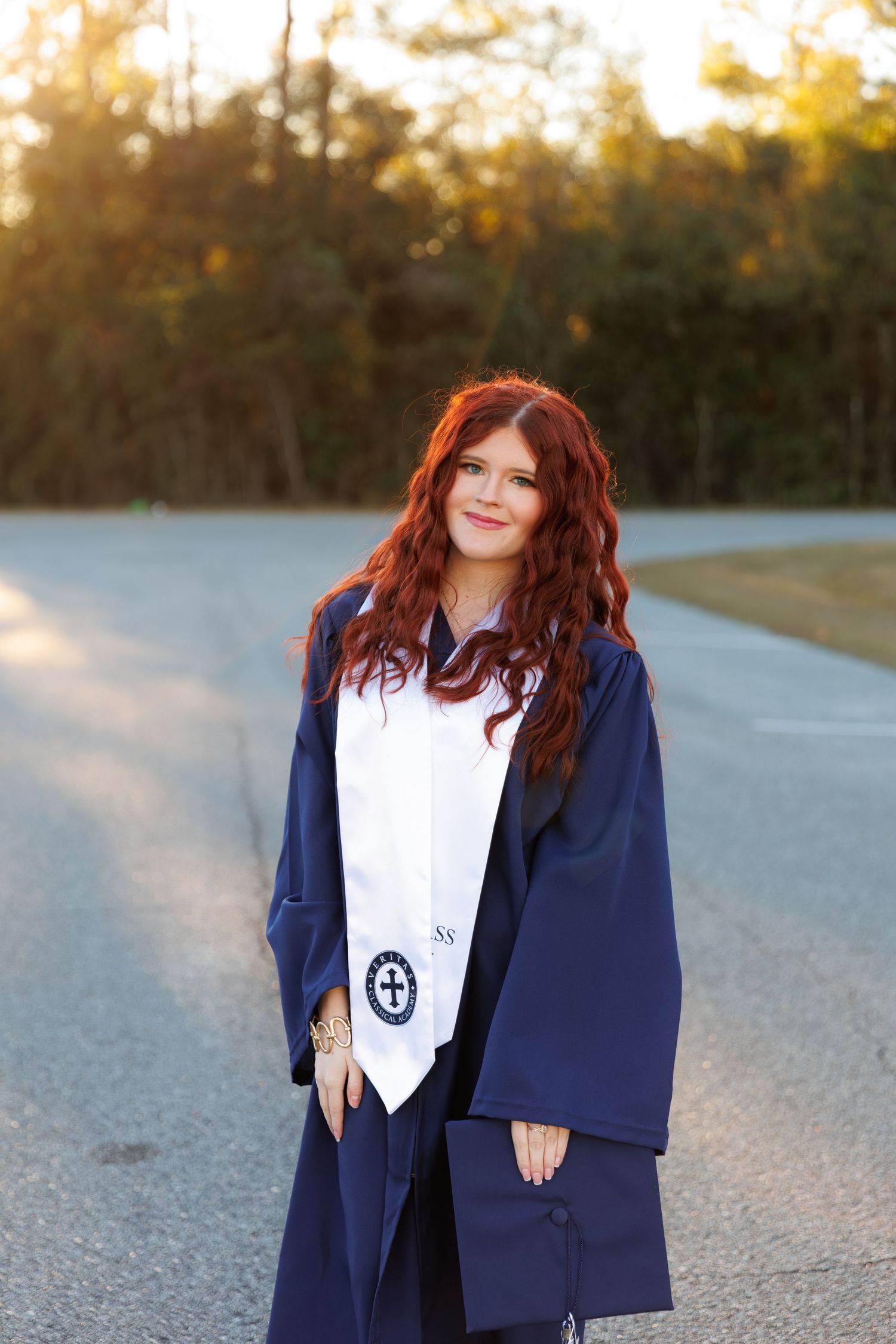 Graduate in navy blue academic regalia and white stole stands on road during golden sunset.