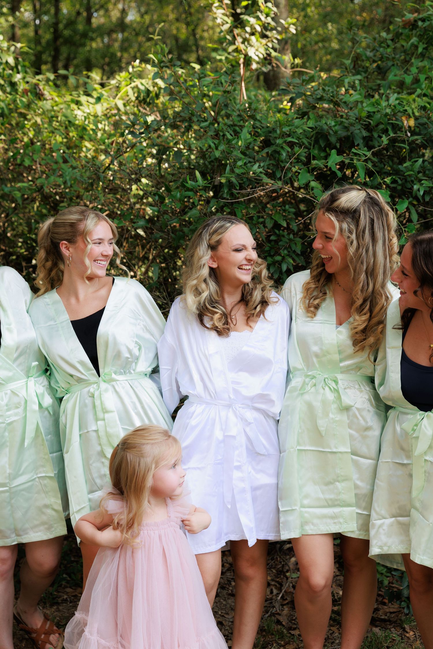A group in matching mint green robes and a little girl in pink stand together laughing outdoors.