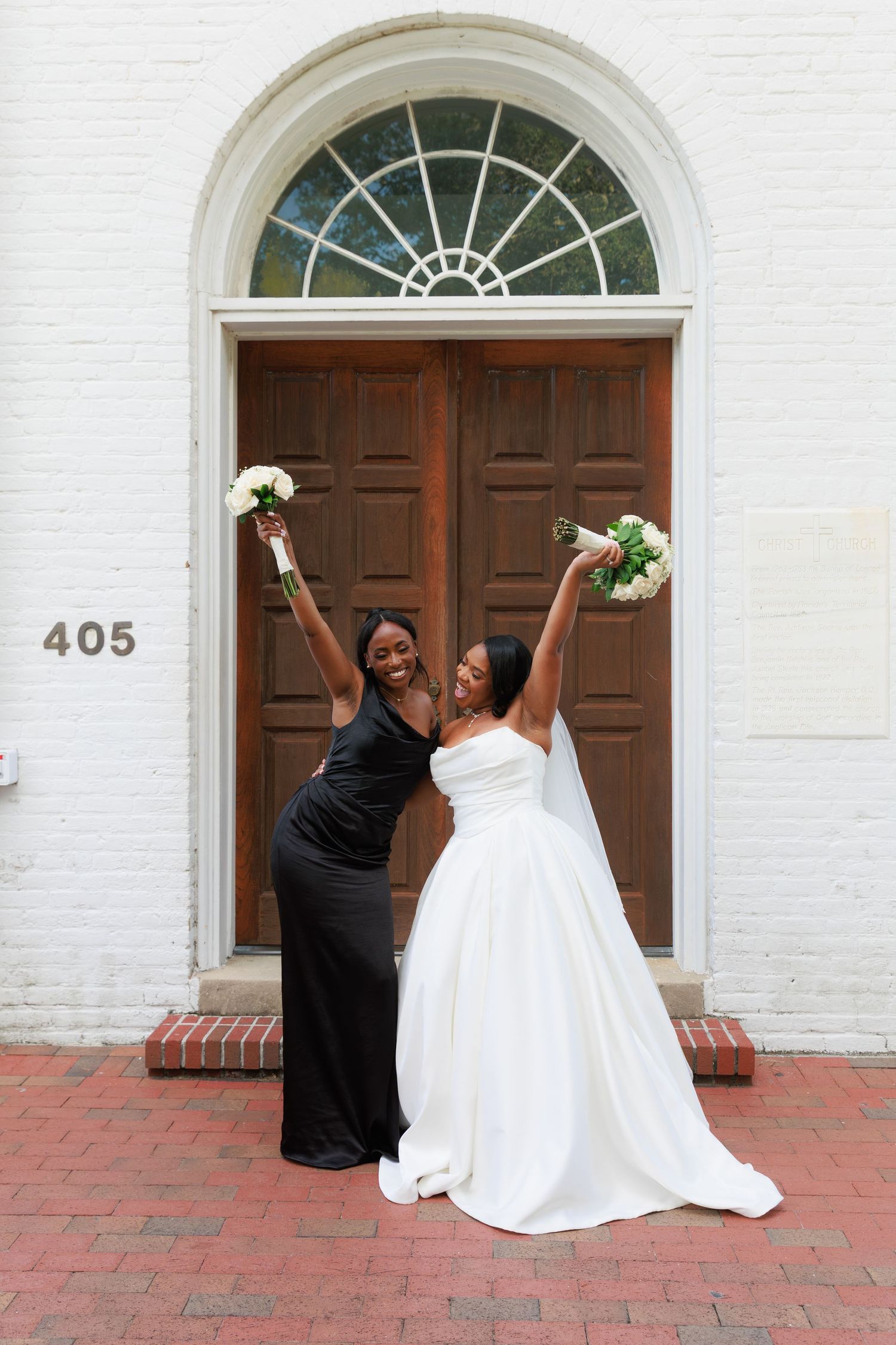 Two wedding attendees in elegant black and white dresses pose joyfully with bouquets in front of a historic doorway.