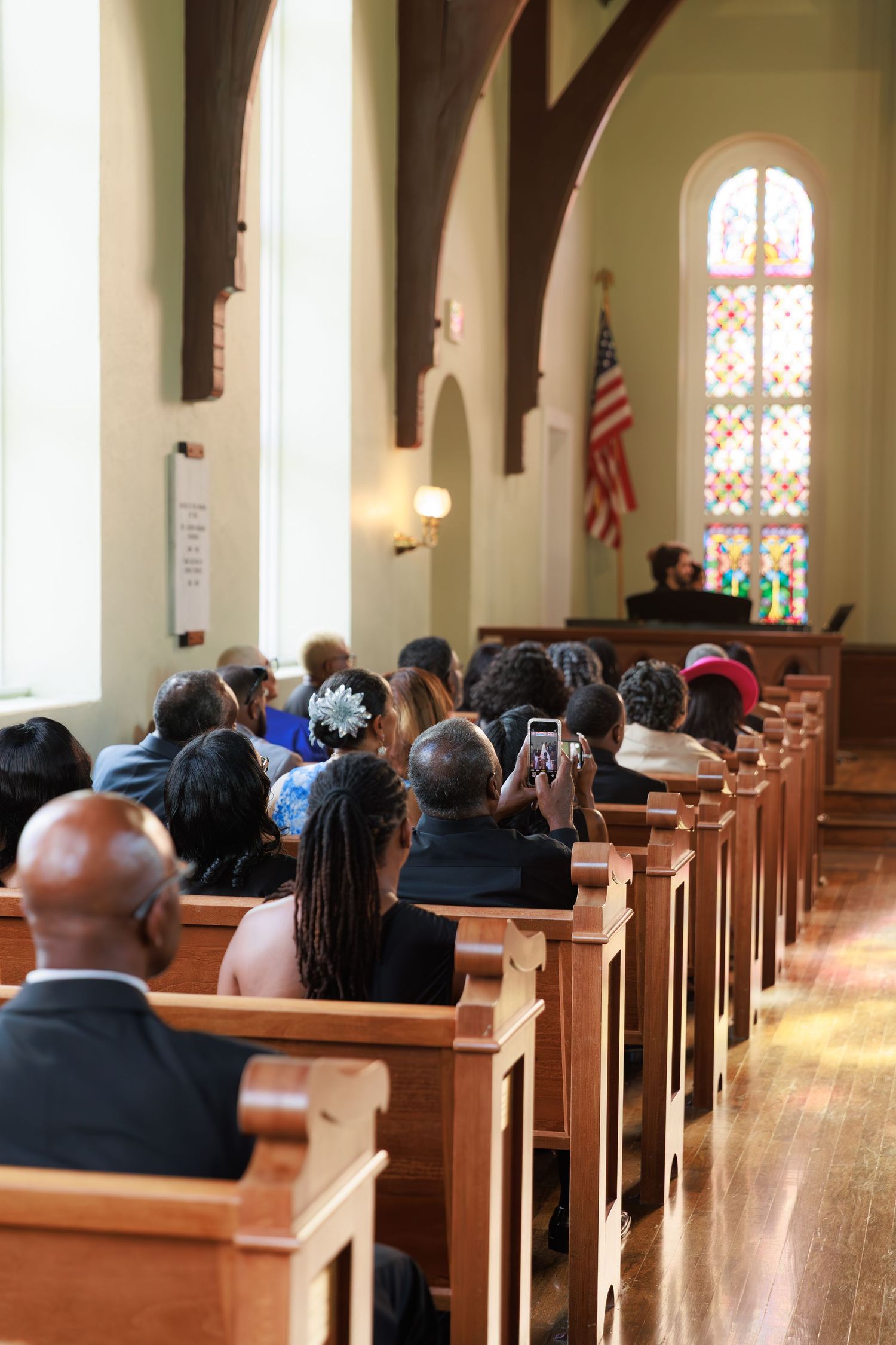 Wedding guests seated in wooden pews at a church ceremony with an American flag and stained glass windows visible.