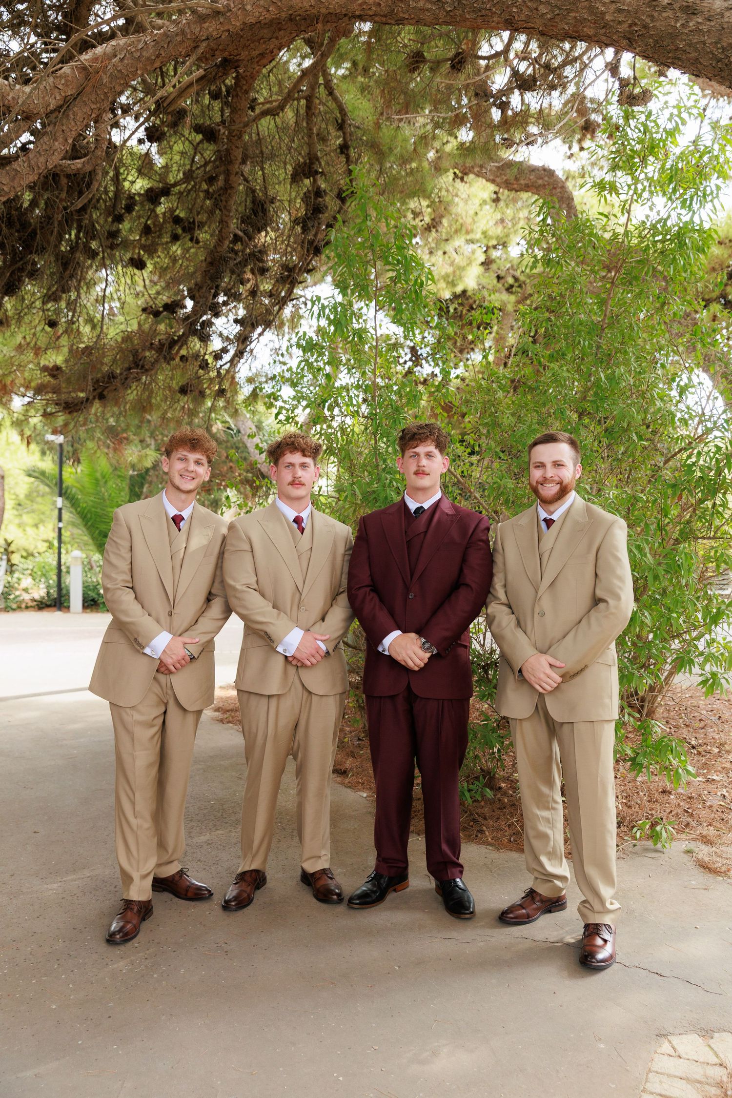 Four men in formal attire stand together under trees in matching tan suits with one wearing burgundy.