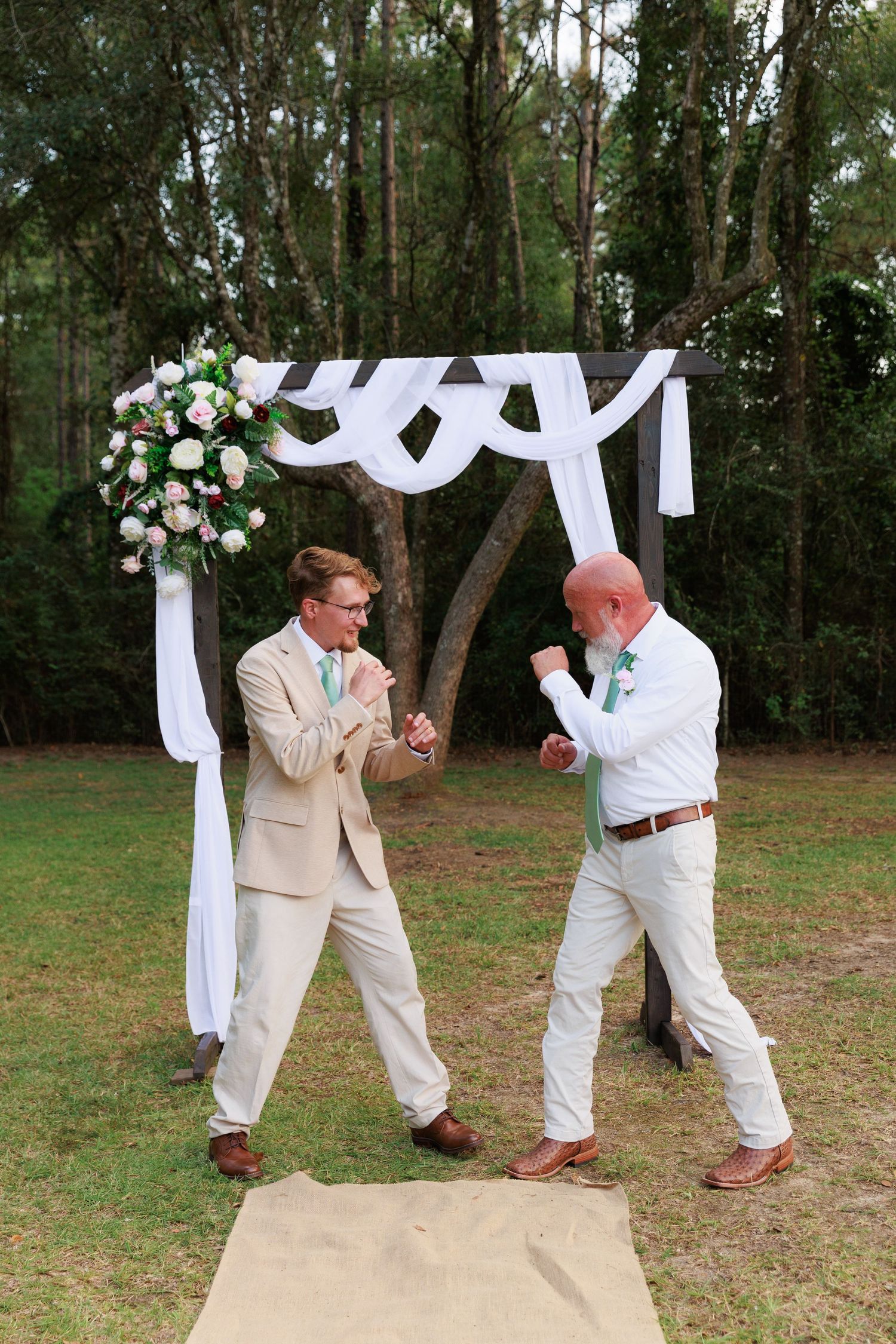 Two people in light colored suits playfully engage in a mock fight sequence outdoors under trees and white draping.
