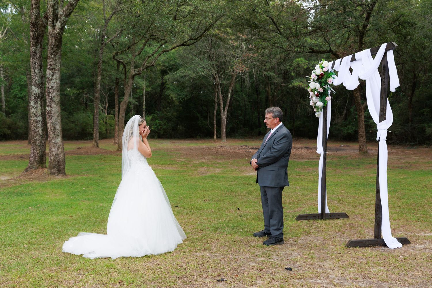 Wedding ceremony taking place in a lush green outdoor setting with white fabric draped between trees.
