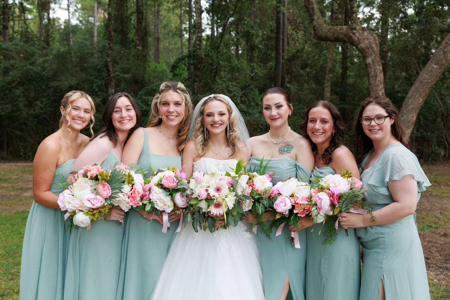 Bridesmaids in seafoam green dresses hold pink and white bouquets while posing with bride in woodland setting.