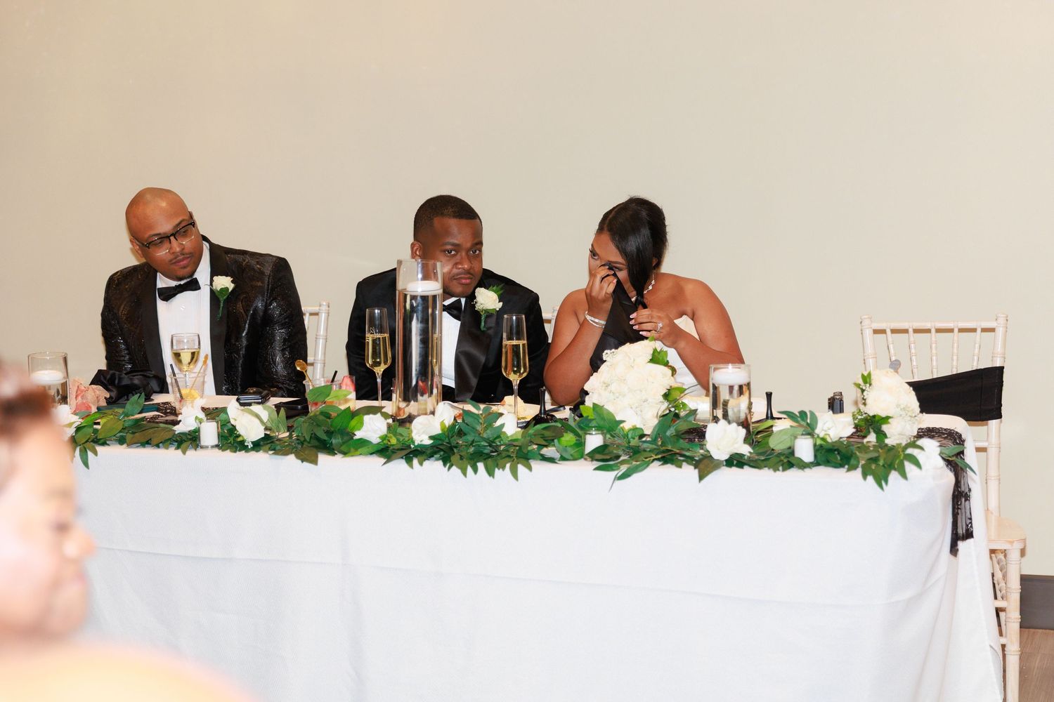 Three guests in formal attire share a candid moment at an elegant head table decorated with white floral arrangements.