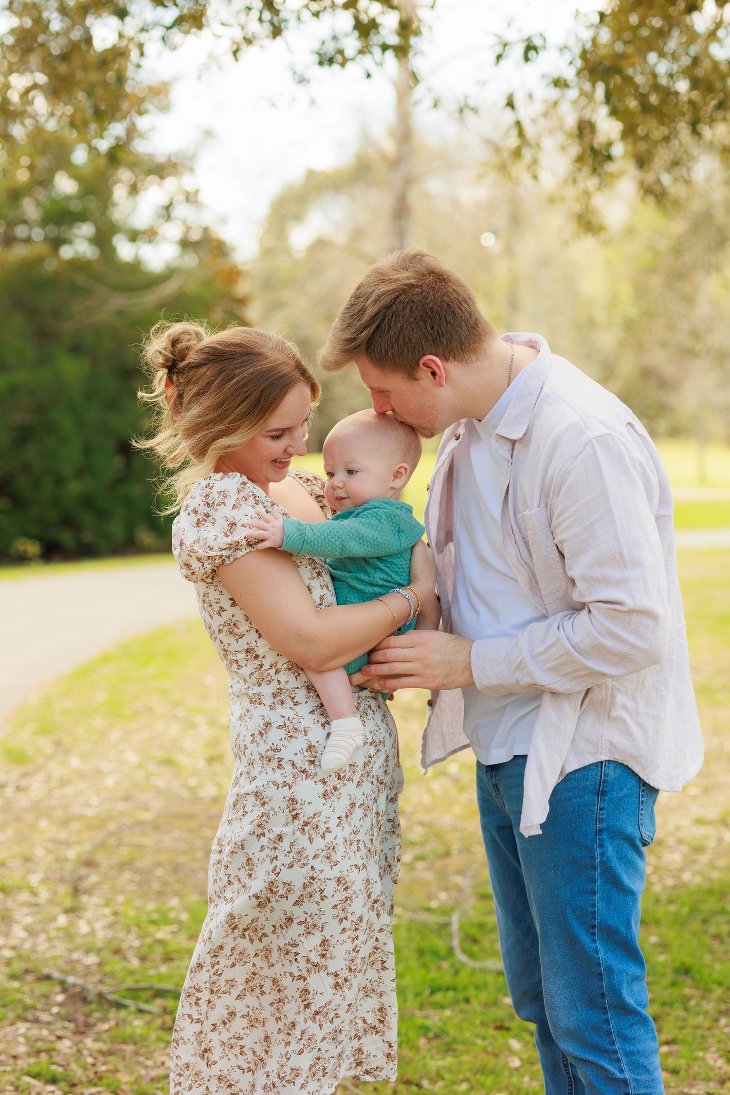 A tender family moment unfolds in a grassy outdoor setting with golden sunlight filtering through trees.