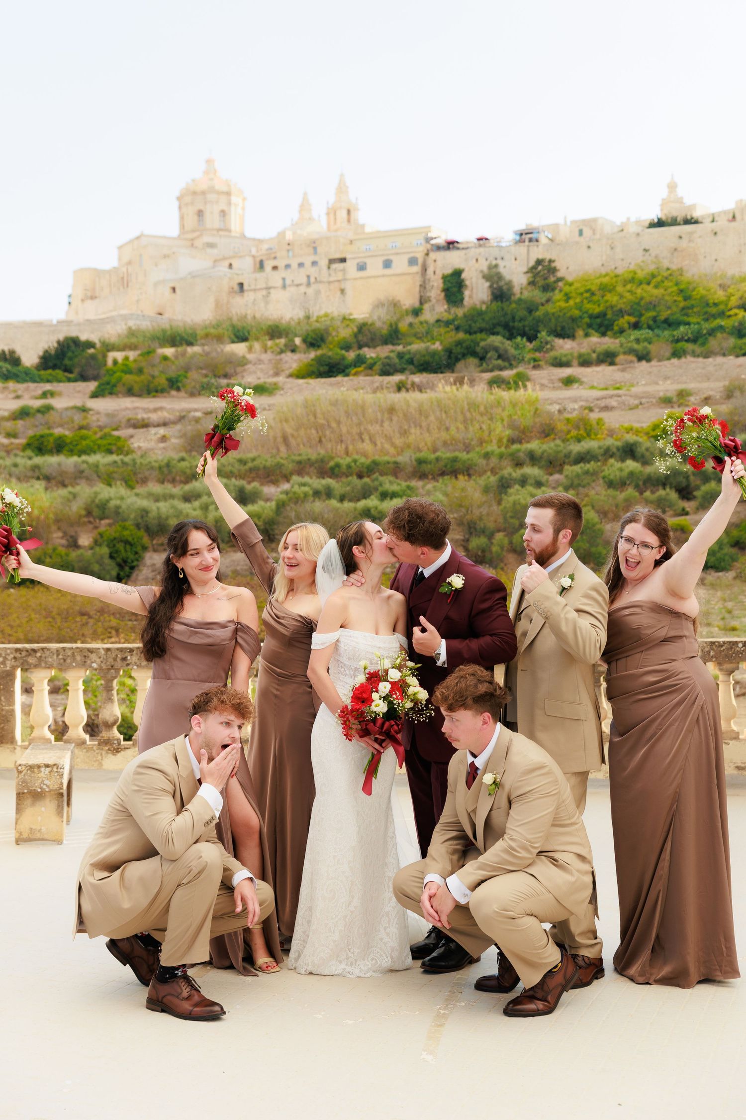 Wedding party poses playfully in front of historic castle with bridesmaids in brown dresses and groomsmen in tan suits.