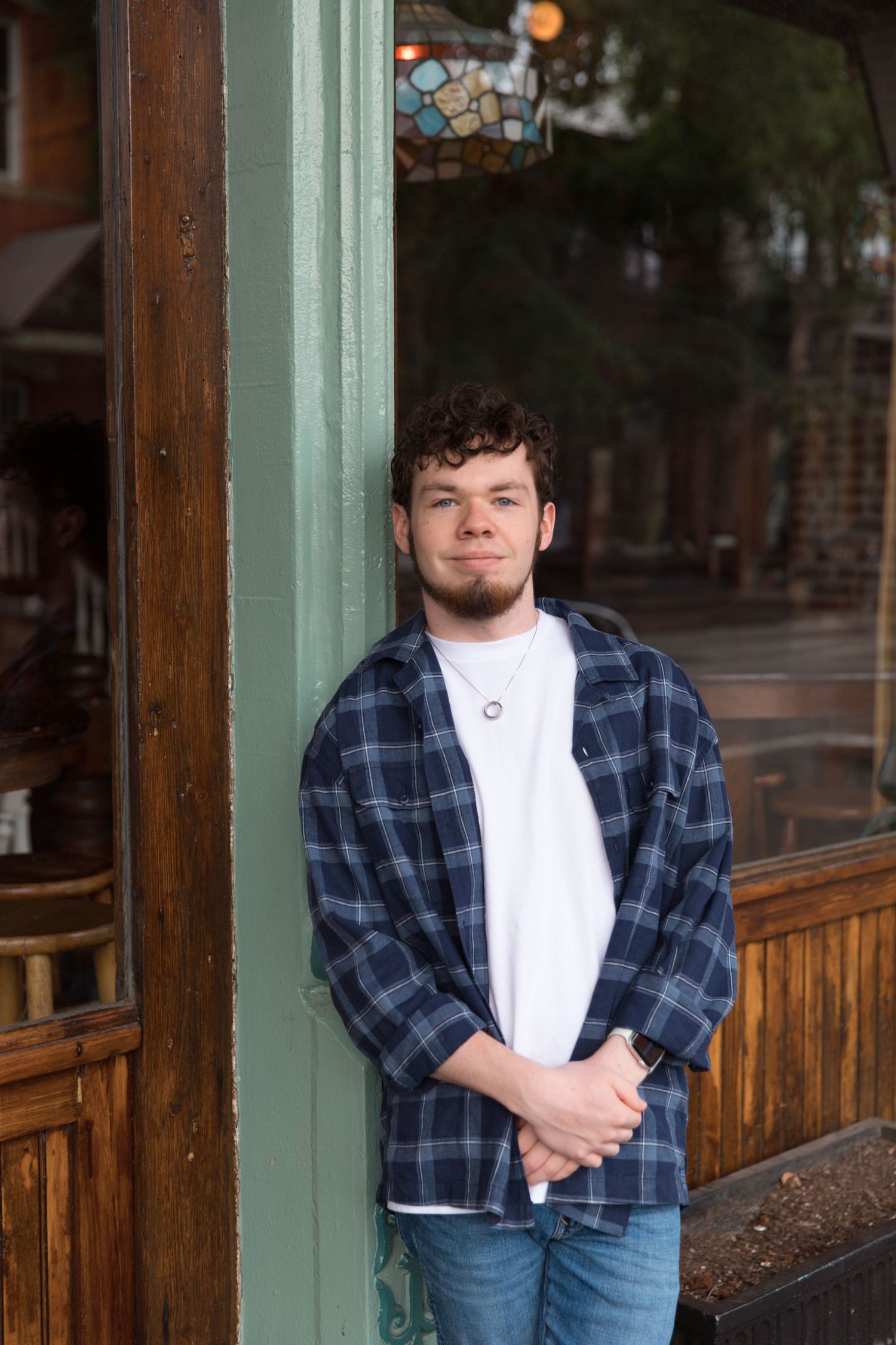 Standing by storefront window in plaid shirt and jeans with casual pose.