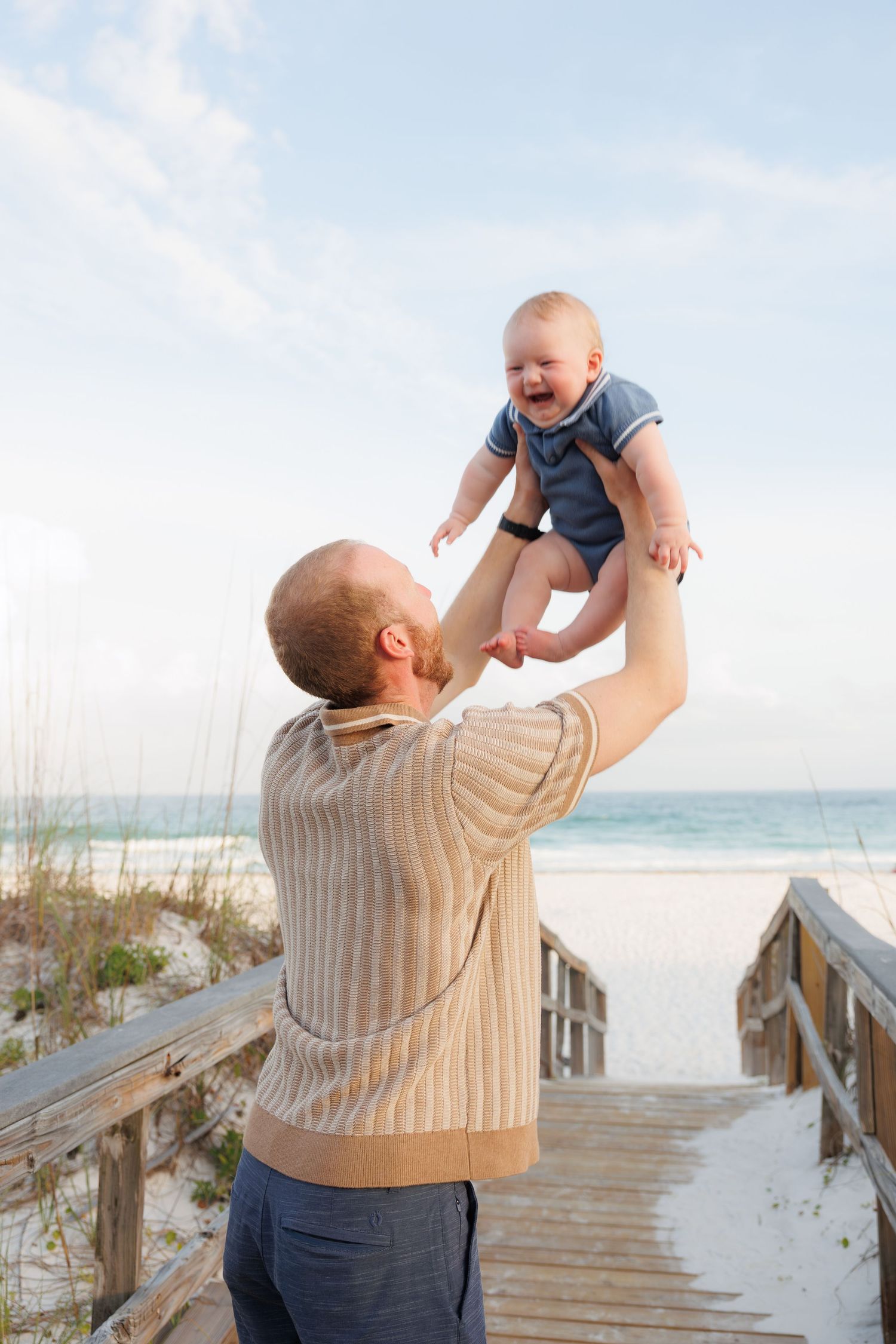 Father lifting smiling baby high in the air on a sunny beach boardwalk with ocean and sand dunes behind.