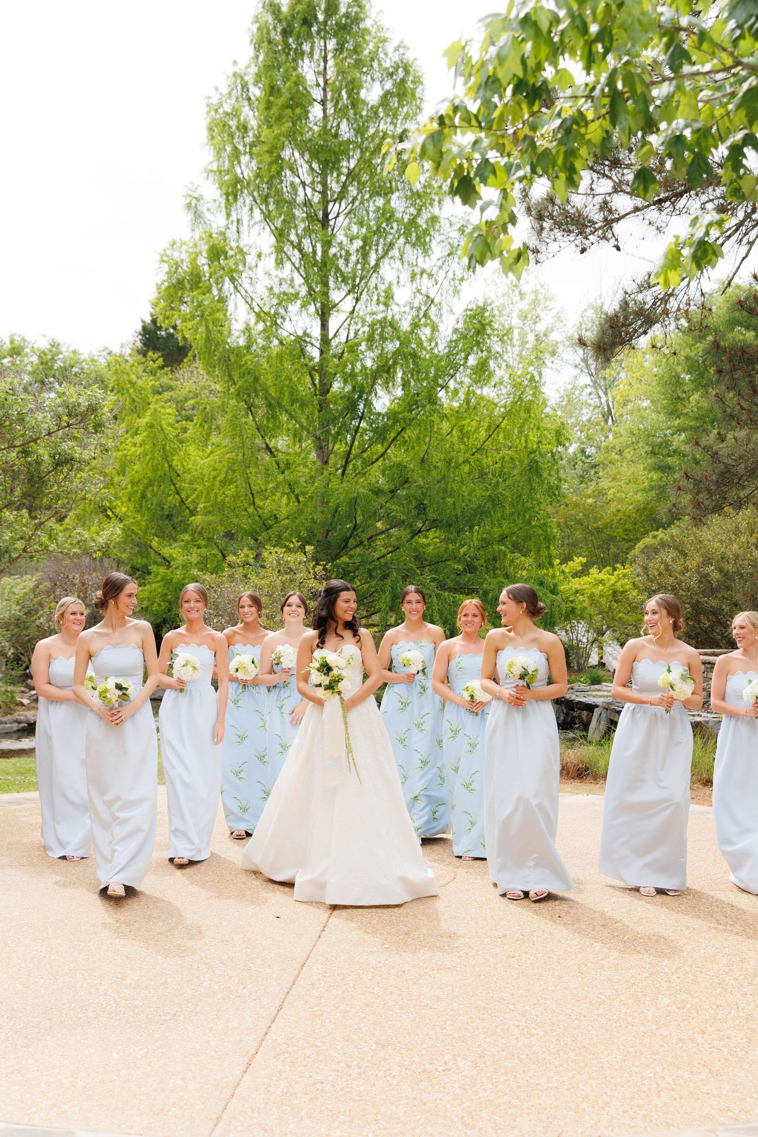 Bride in white gown walks with bridesmaids in light blue dresses holding white bouquets outdoors.