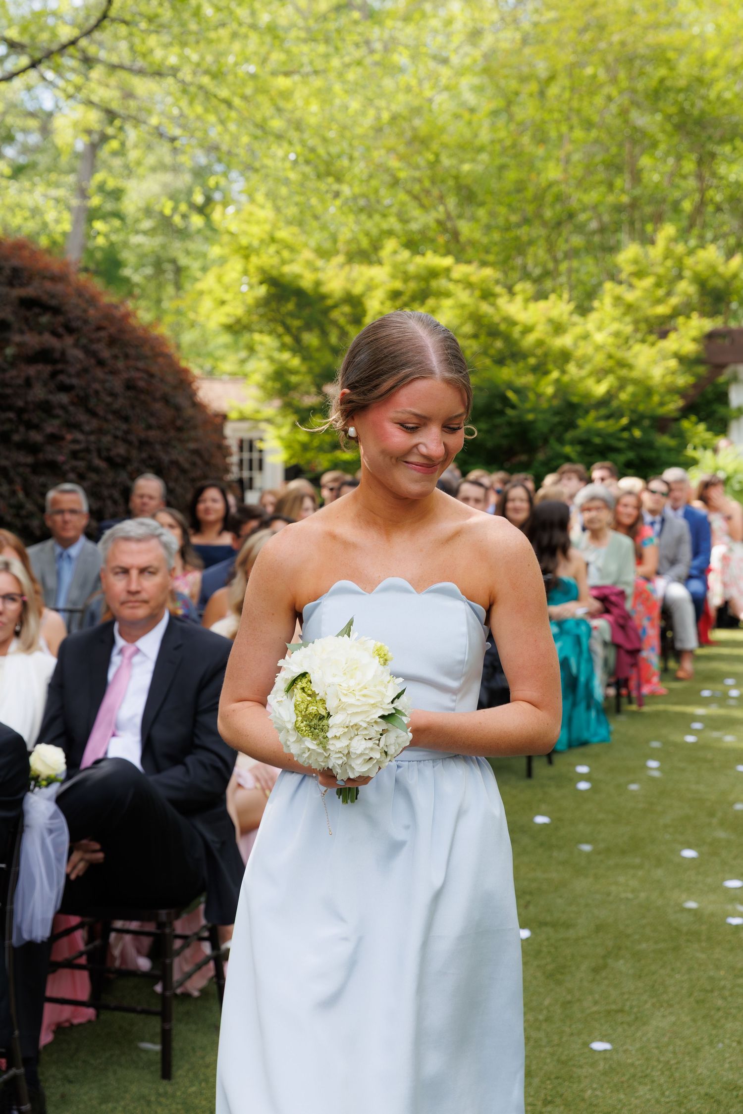Smiling bridesmaid in light blue strapless gown walks down outdoor wedding aisle holding white floral bouquet.