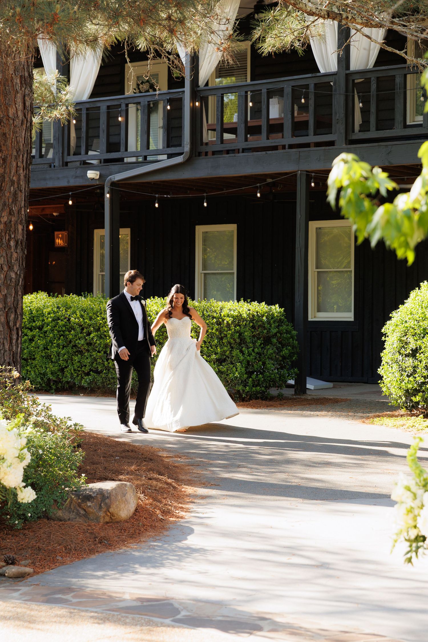 Bride and groom walk hand in hand along a path outside a elegant dark-wood venue surrounded by lush greenery.