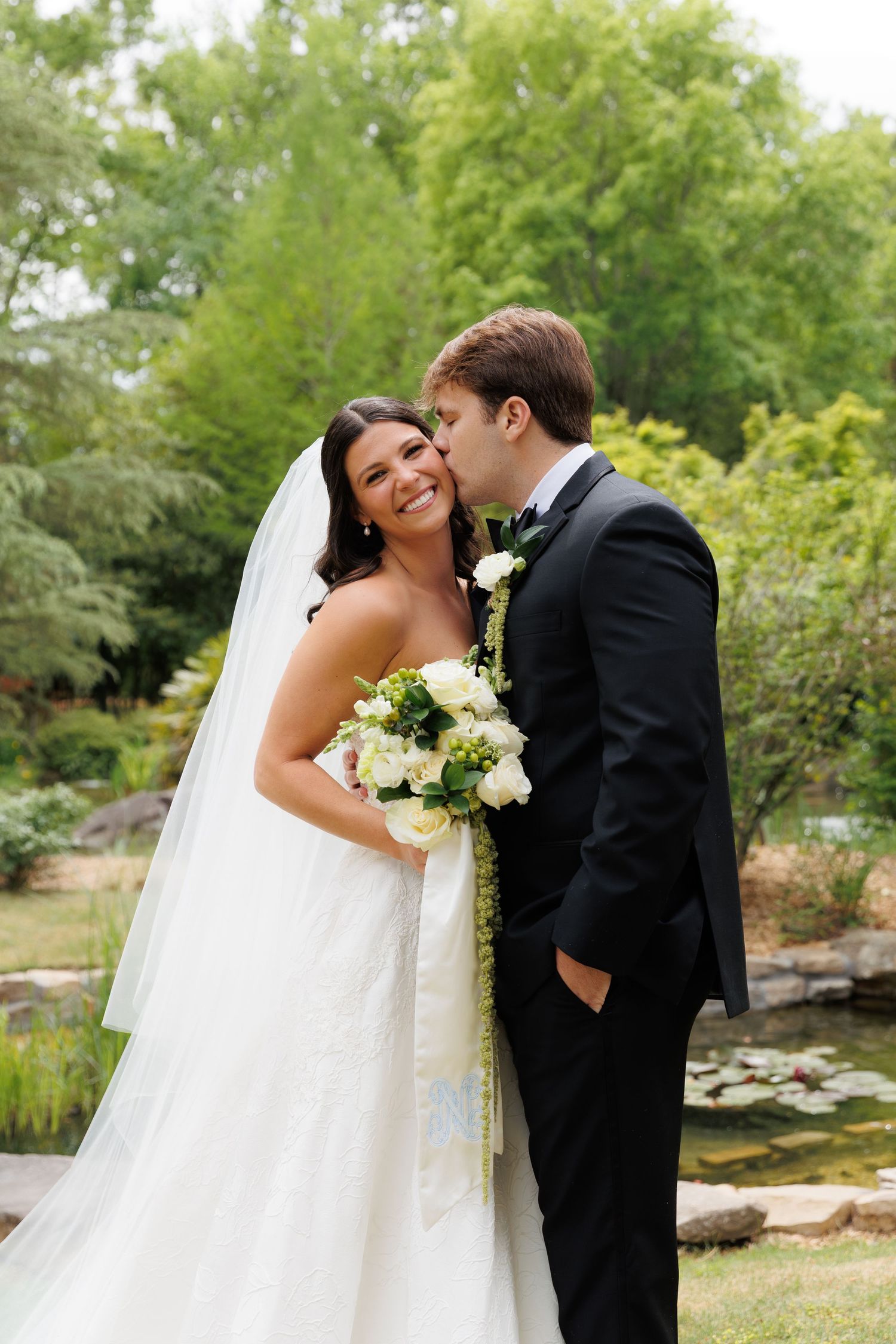 Groom kissing smiling bride on cheek outdoors, bride holding white bouquet in garden.