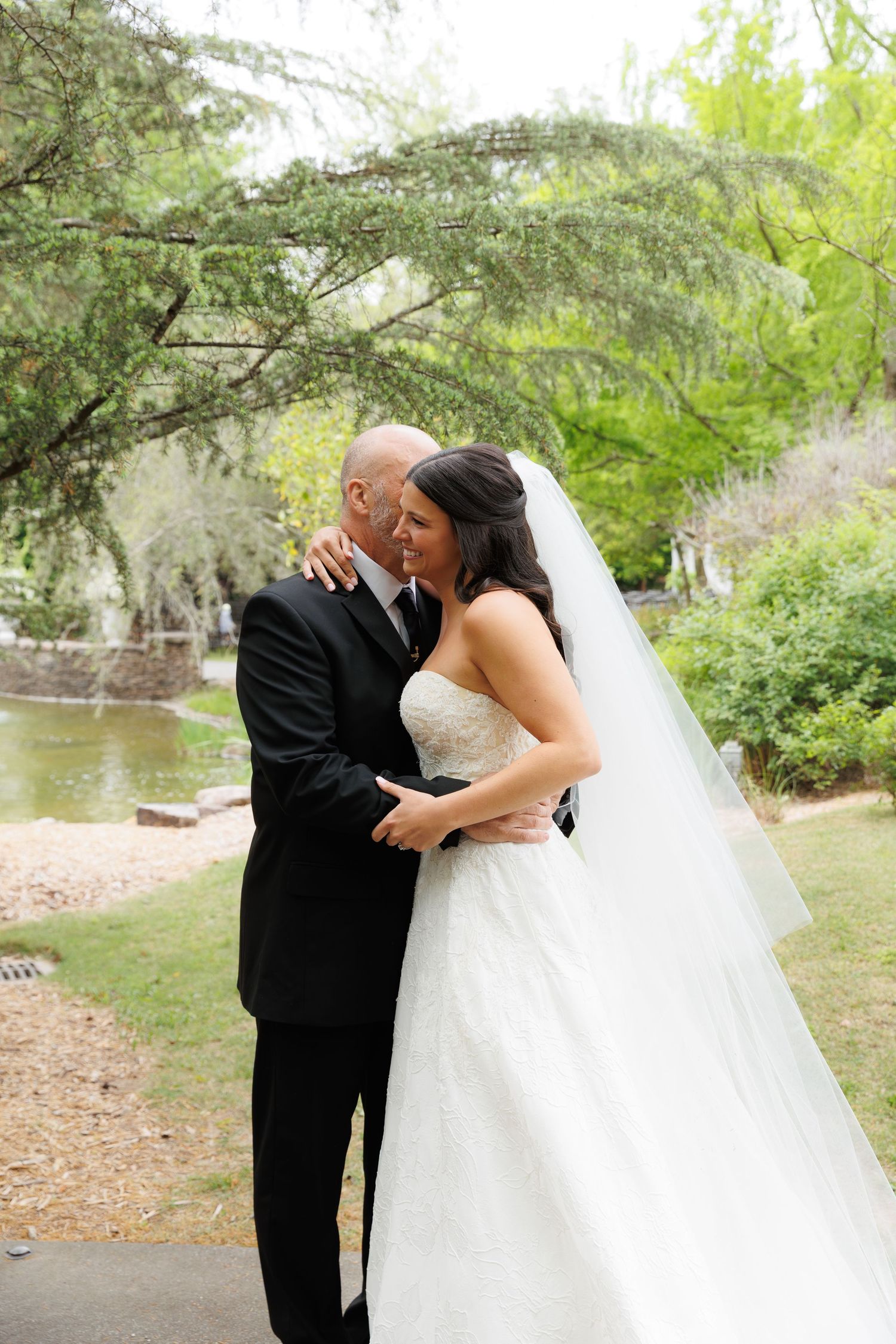 Bride and groom share an intimate embrace in a lush garden setting, with the bride in a white gown and veil.
