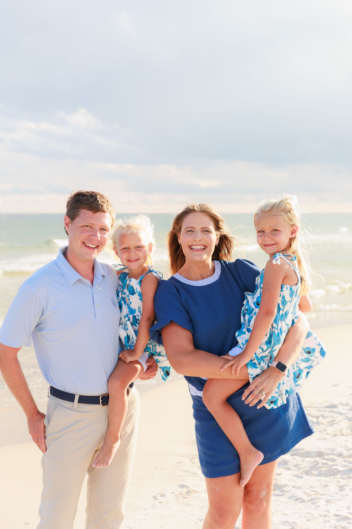 Happy family of four posing on a sunny beach, parents holding two young daughters in matching blue floral dresses.