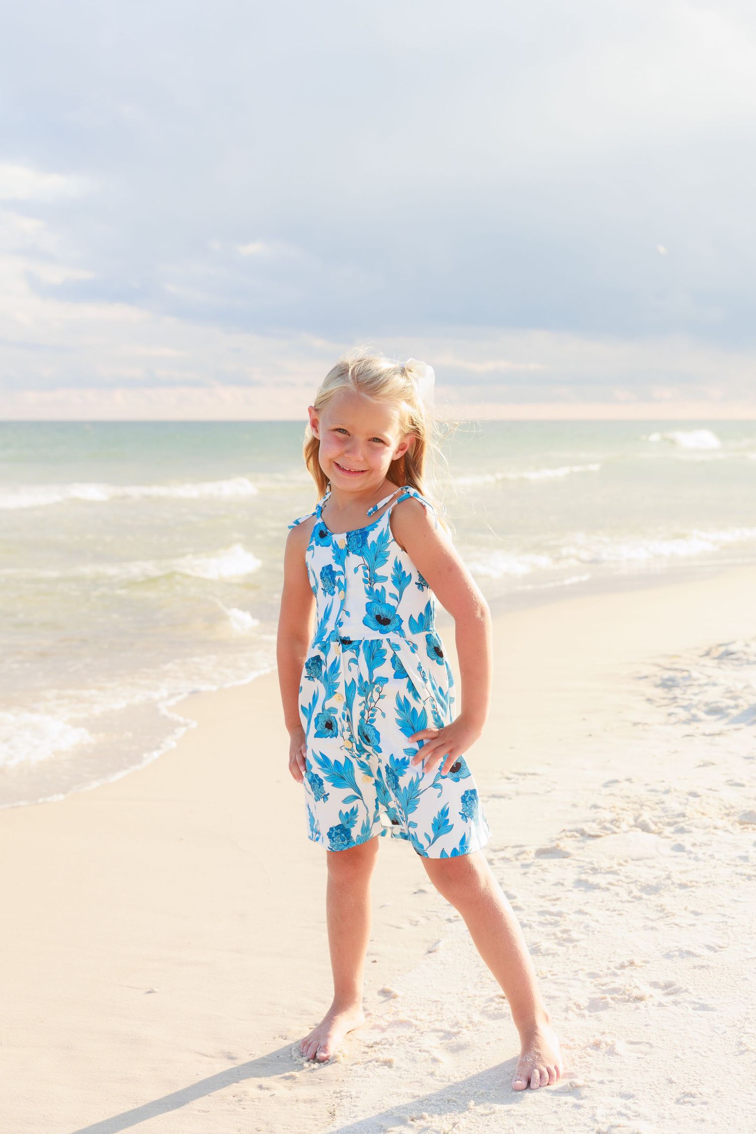 Happy young blonde girl in blue floral romper smiling and posing on a sunny white sand beach.
