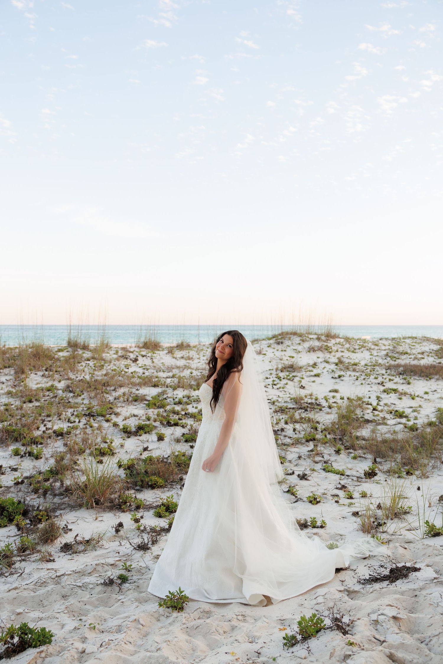 Bride in strapless white gown and cathedral veil poses on white sand beach with turquoise ocean backdrop.