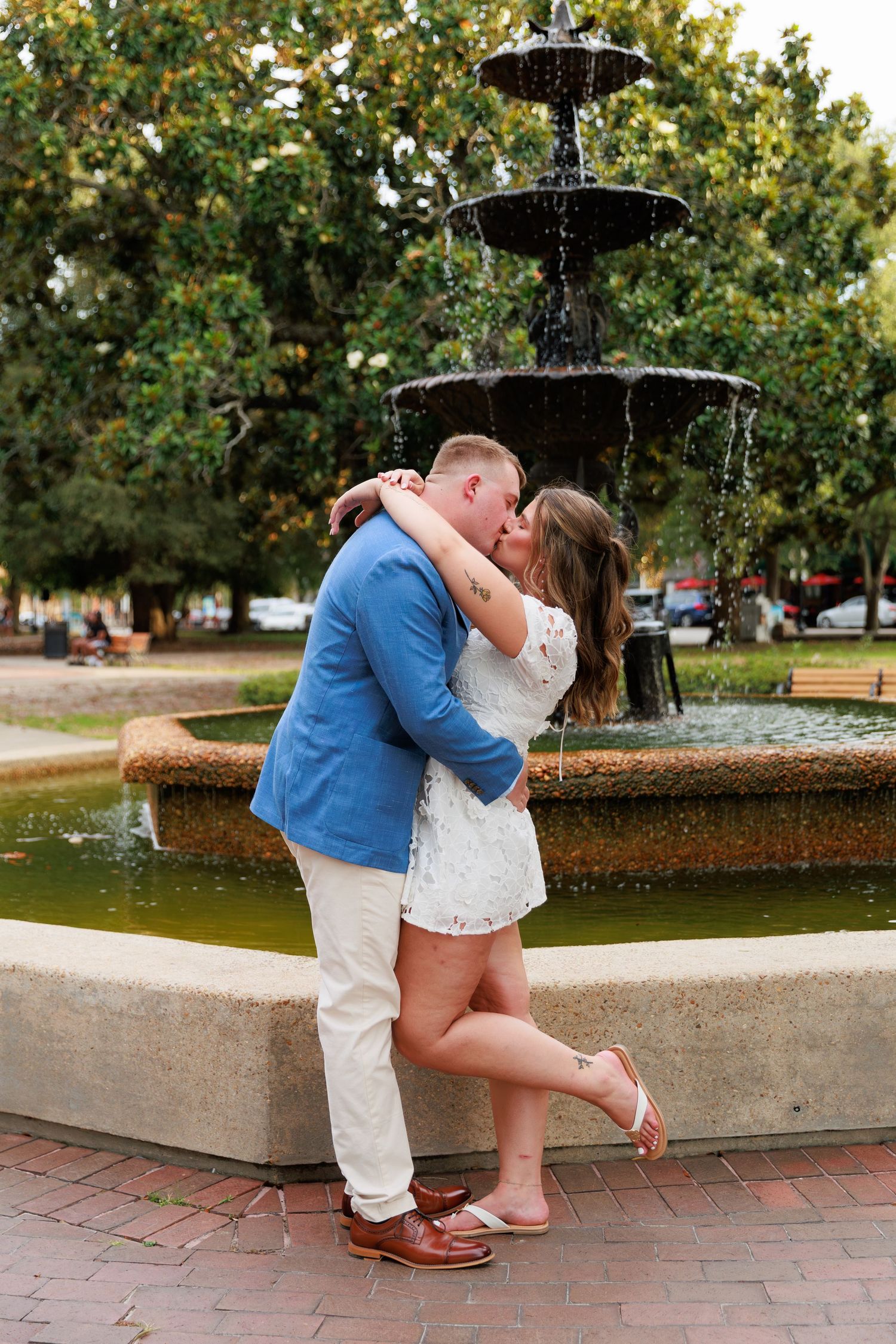 Couple sharing a romantic kiss in front of a tiered stone fountain in a lush green park.