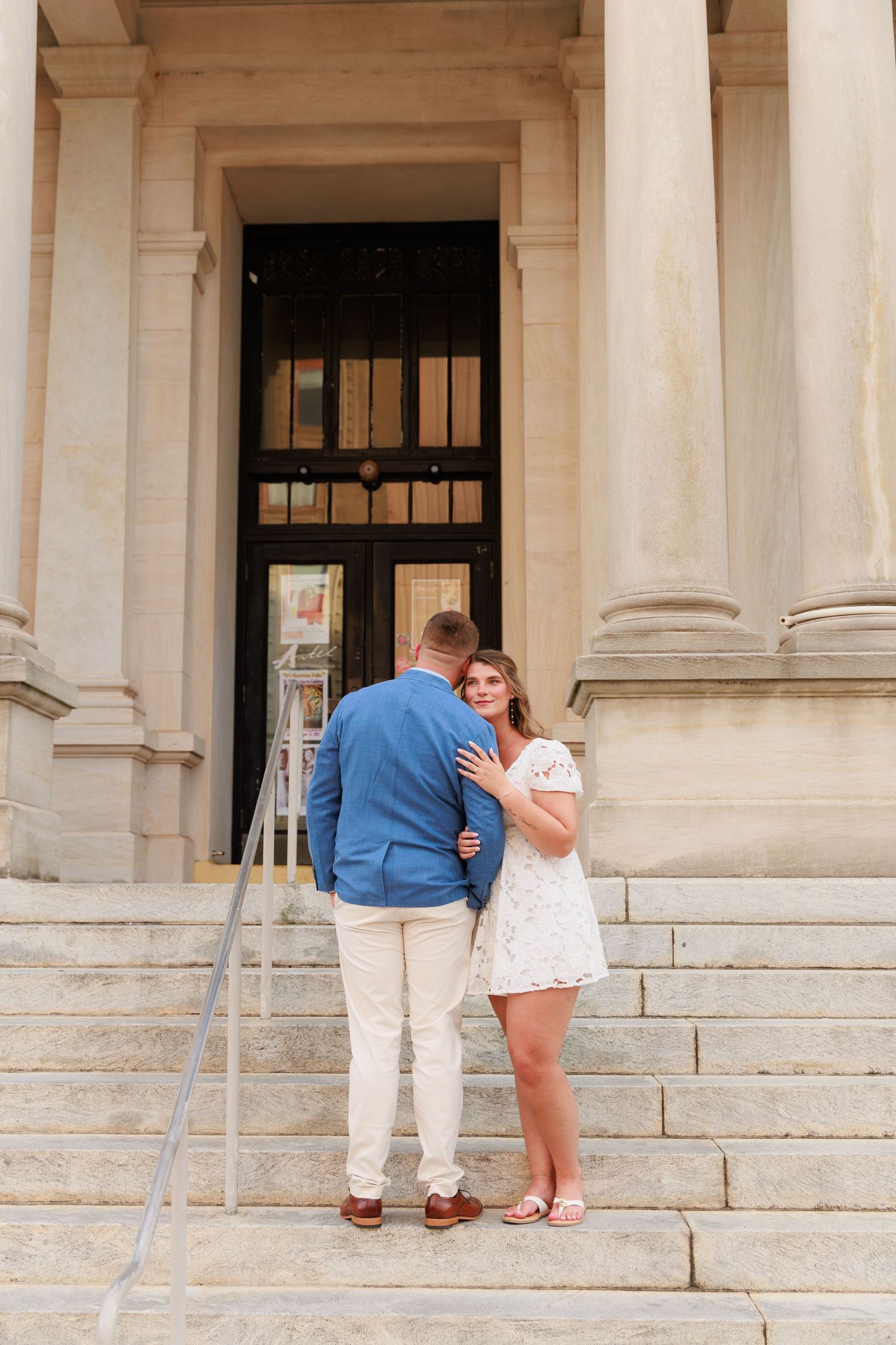 Couple embracing on stone steps of a classical building, woman in white dress smiling at camera, man in blue blazer.