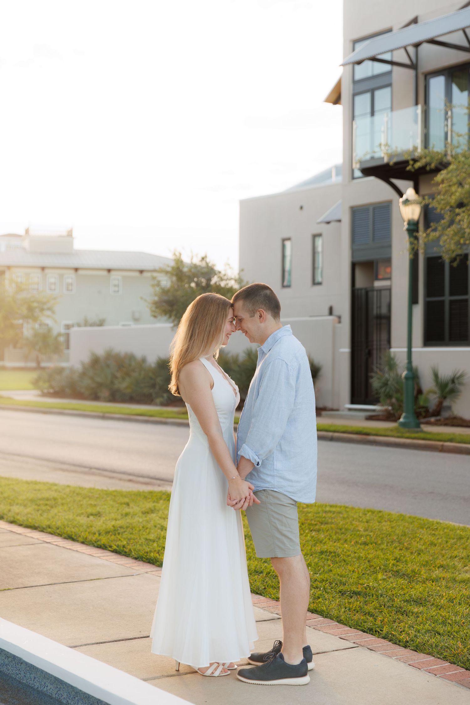 Couple holding hands and touching foreheads on a sidewalk at sunset near a modern white building.