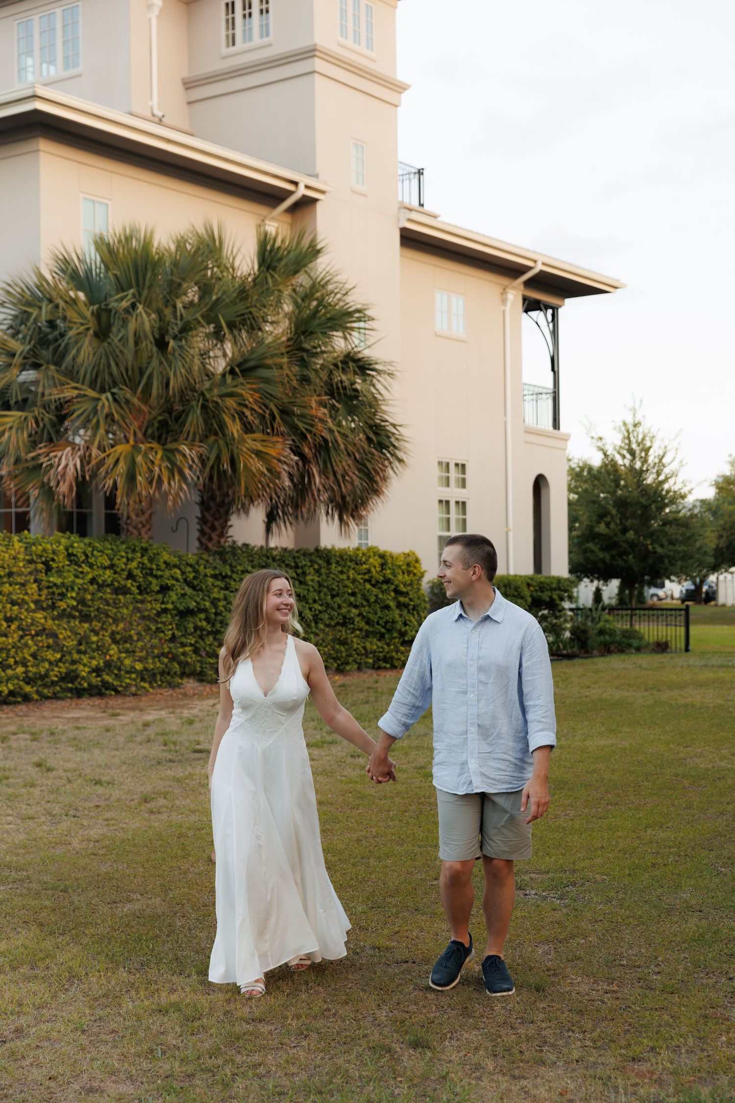 Smiling couple holding hands and walking on grass in front of a large building with palm trees.