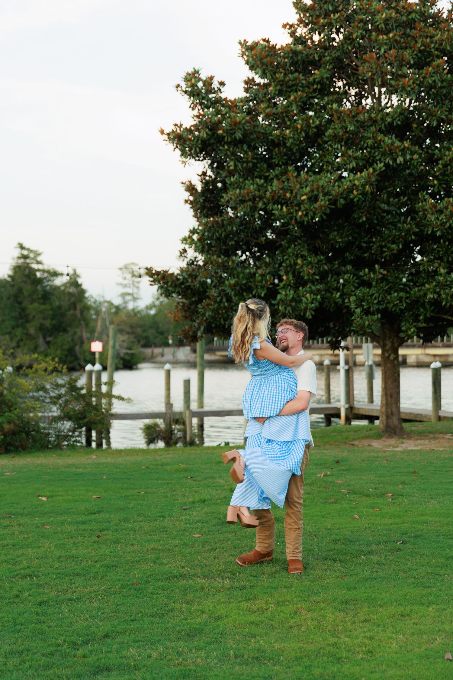 A man lifts a woman in a blue dress while embracing outdoors near a waterfront with lush trees.