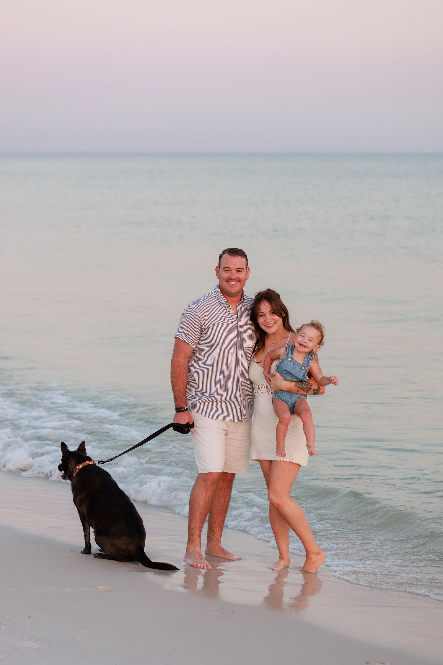 Family with a baby and black dog posing on a sandy beach at sunset with calm turquoise ocean waves.