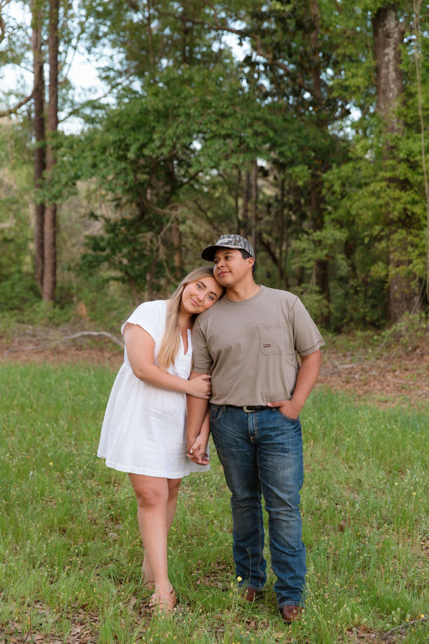 A young couple poses in a grassy field, she in a white dress leaning on him in a tan shirt and jeans.