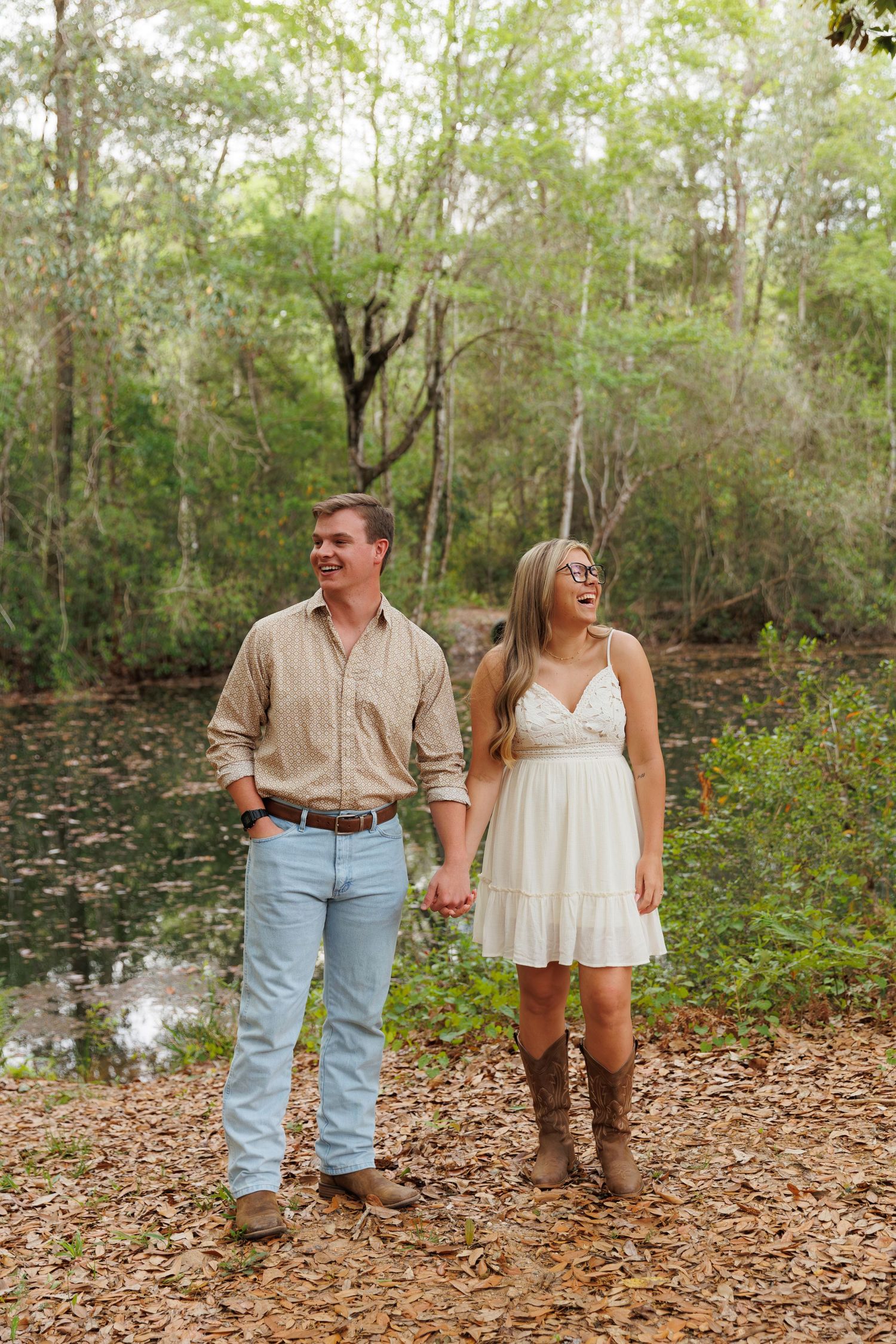 Couple holding hands walking near a pond in a lush forest, woman in white dress and boots, man in jeans and tan shirt.