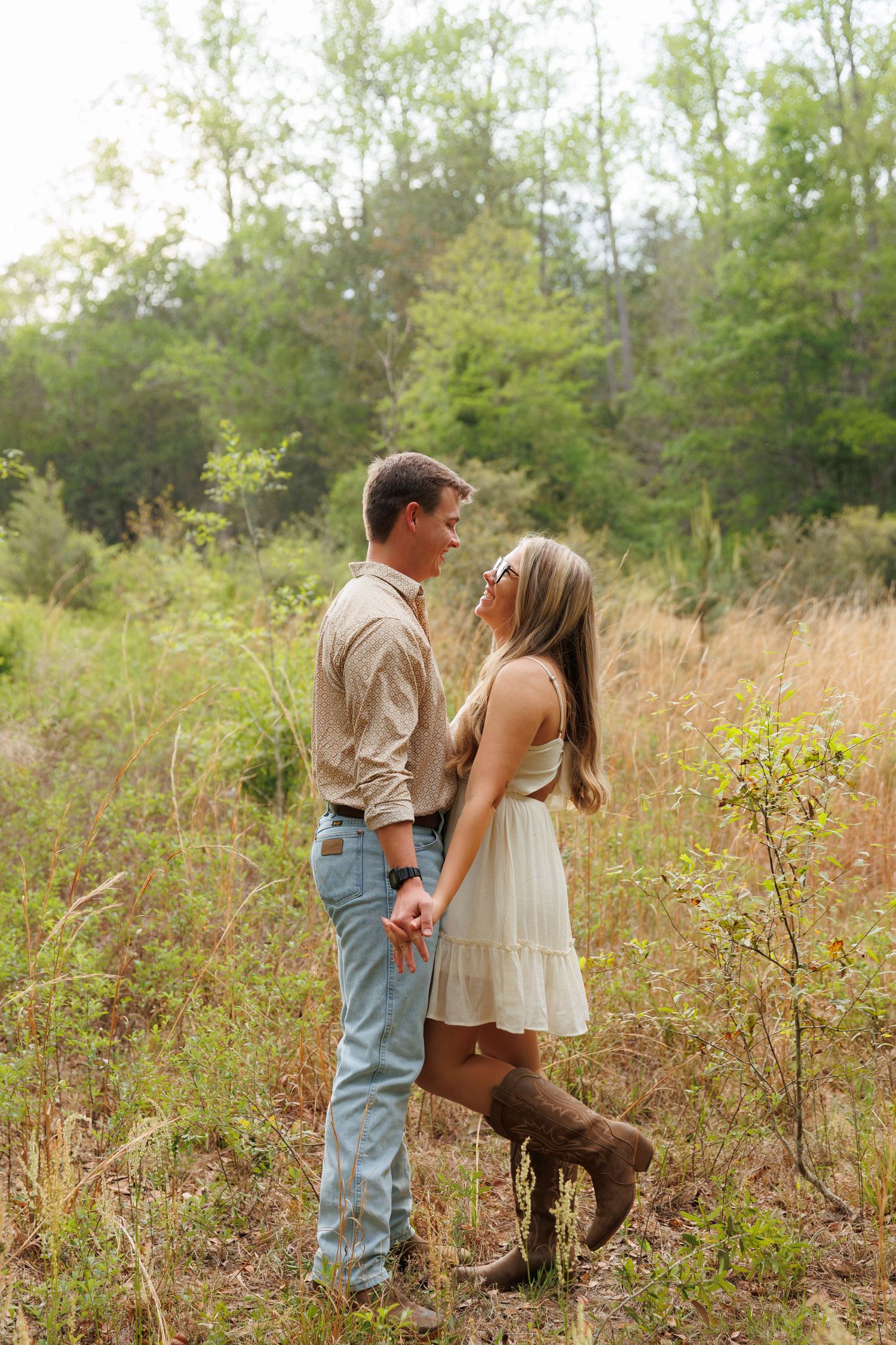 Couple holding hands in a rustic field, woman in white dress and boots, man in jeans and beige shirt, surrounded by greenery.