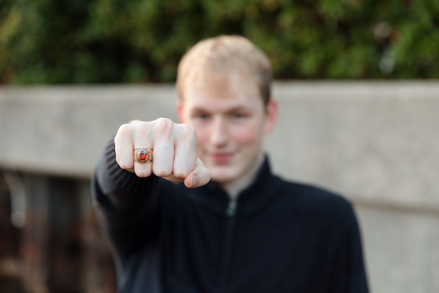 Young blond man in black jacket extending fist toward camera, wearing a red gemstone ring, with blurred background.