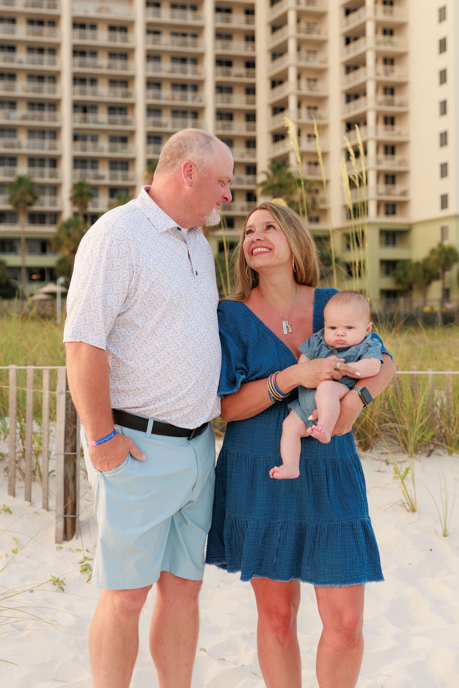Couple with baby smiles on sandy beach in front of resort hotel during golden hour.