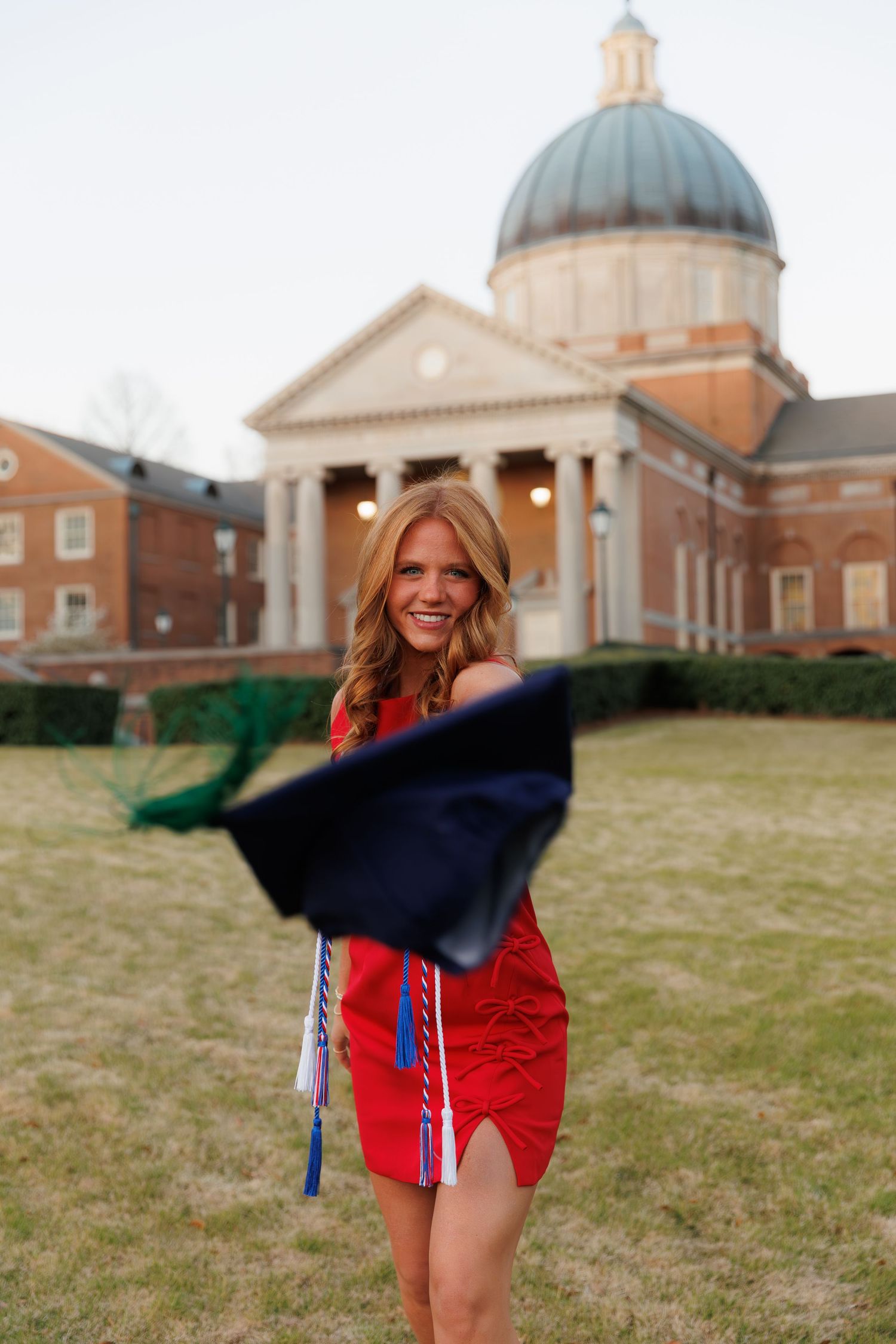 Graduate in red dress tosses navy cap in front of domed university building.