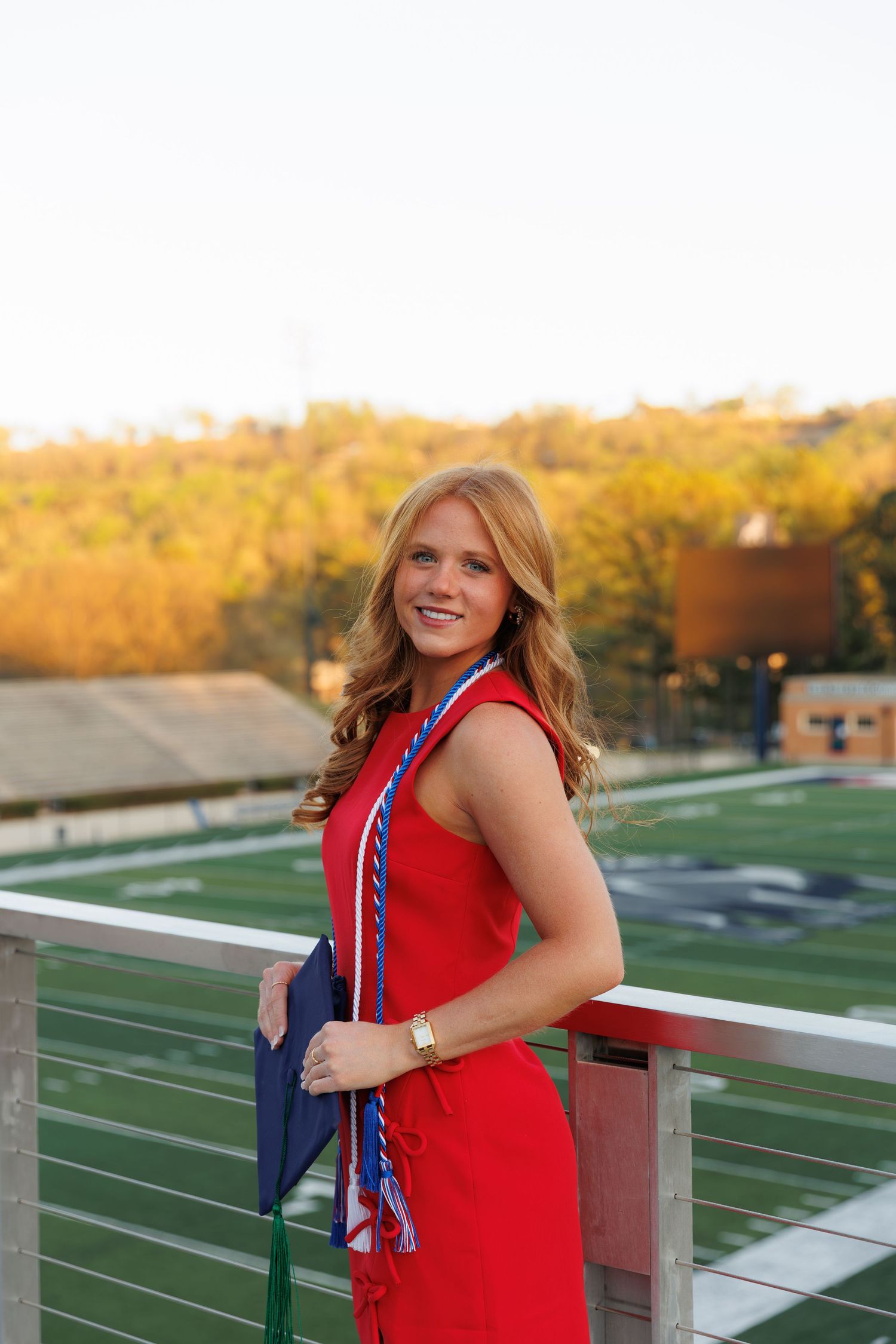 Young woman in red dress holding graduation cap with colorful cords, posing on stadium bleachers at sunset.