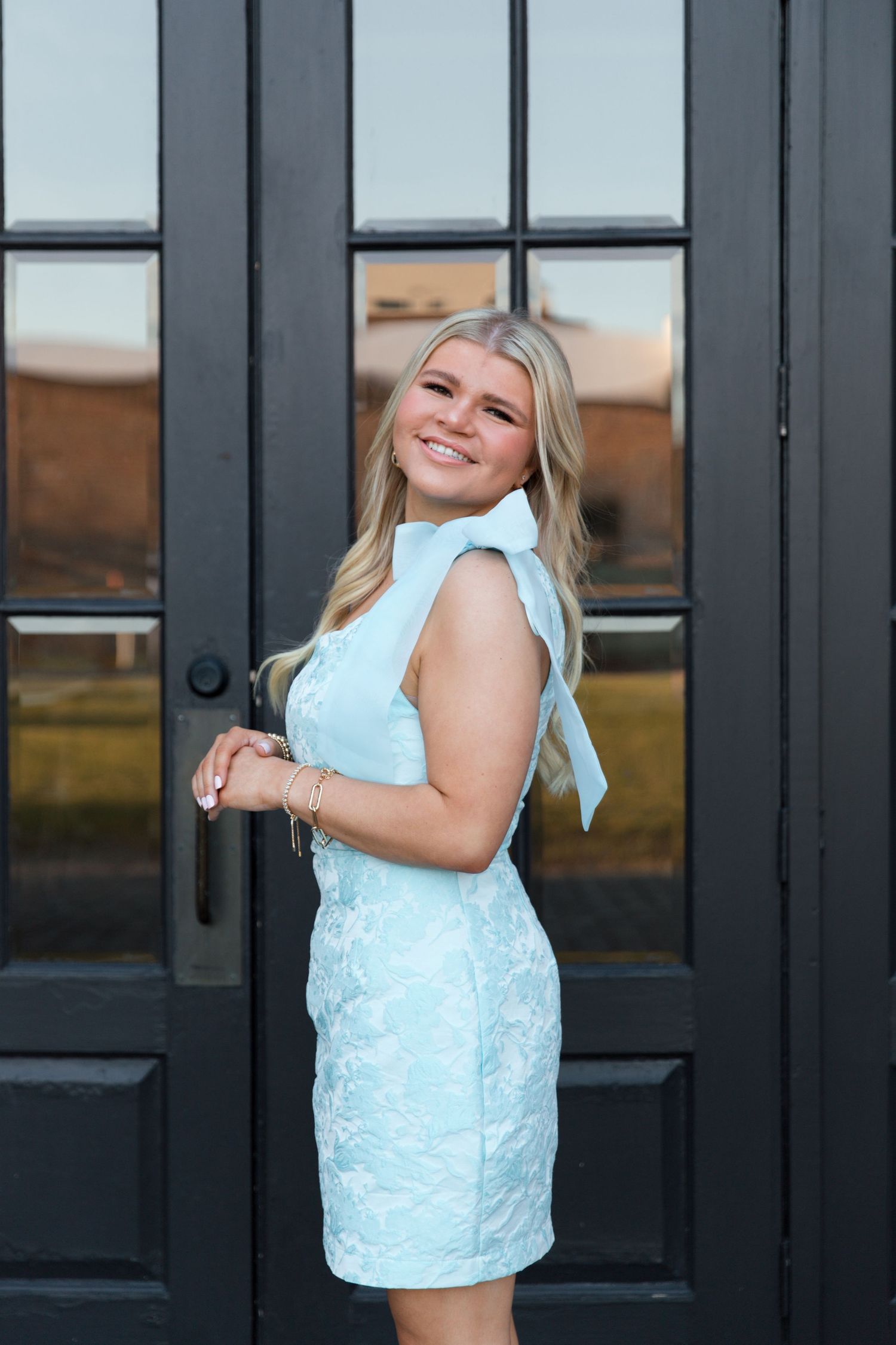 A woman in a white dress poses playfully against black doors during a photo shoot.