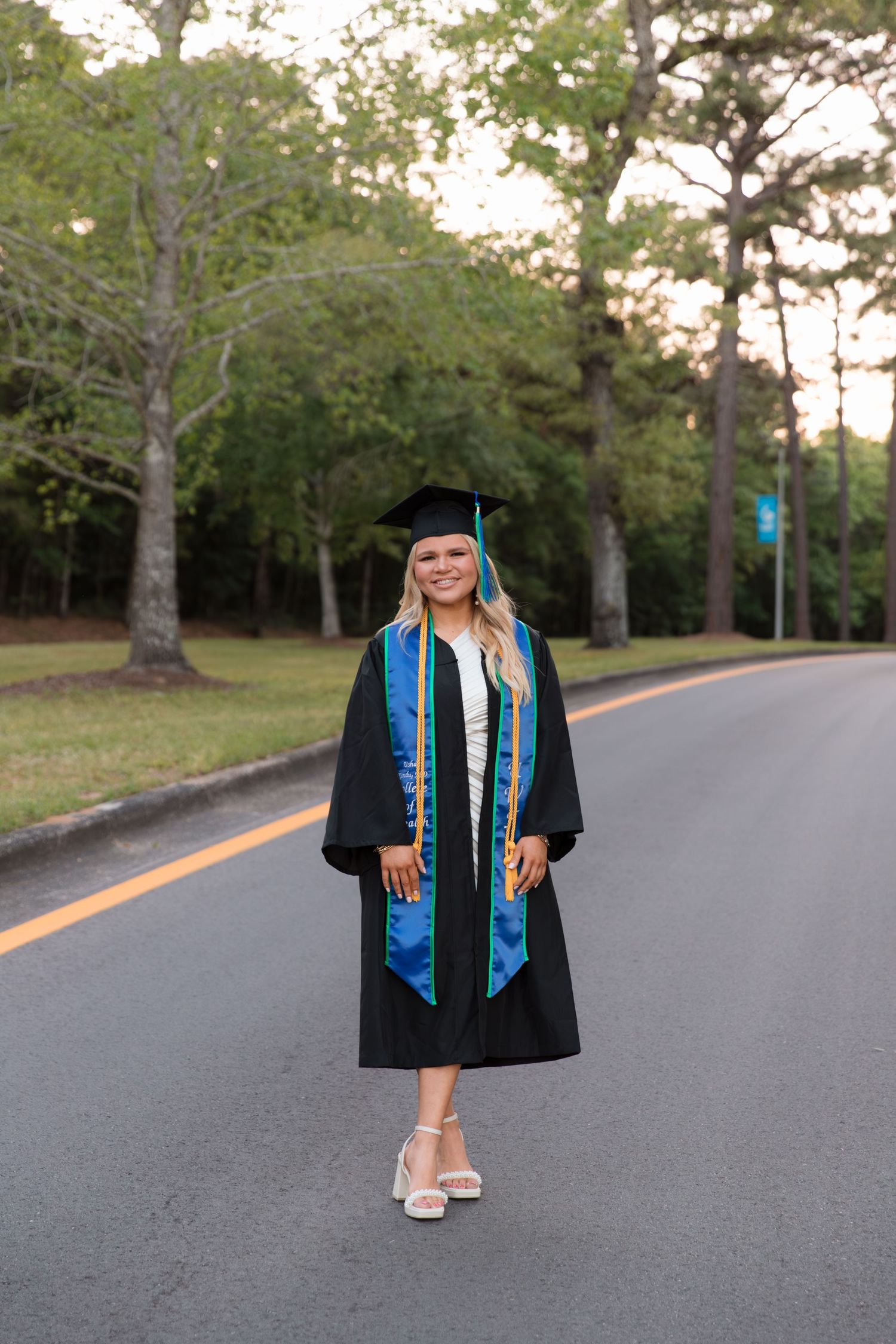 Graduate in academic regalia poses confidently on a tree-lined campus street at sunset.