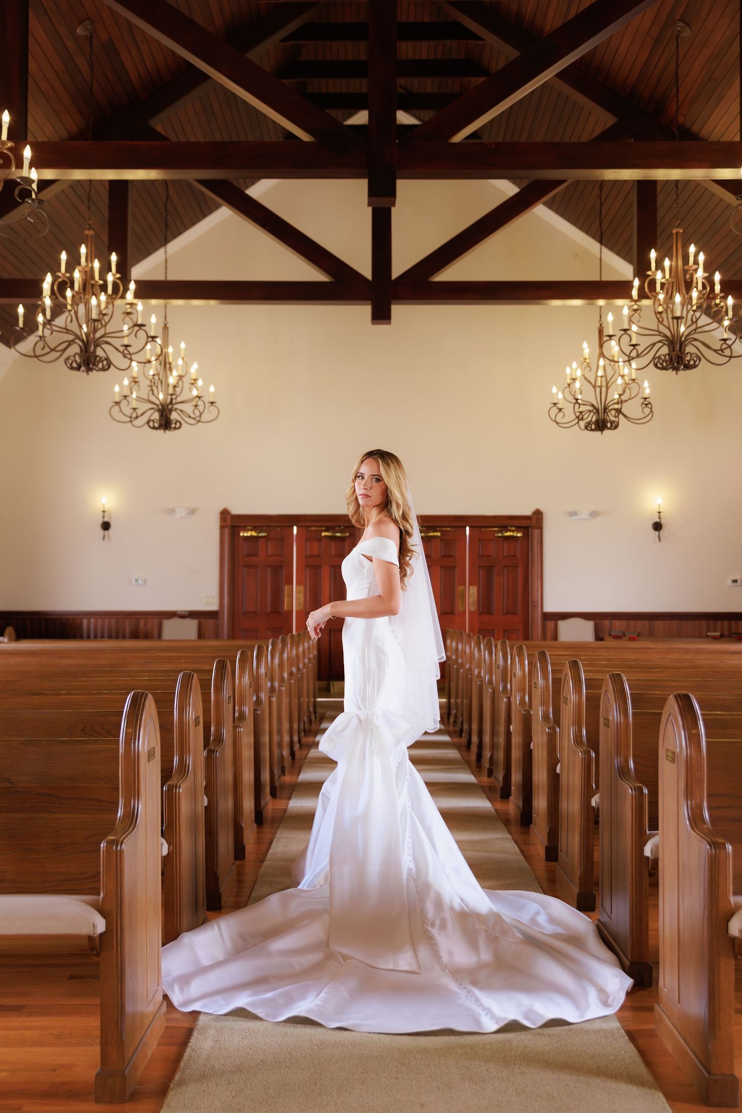 A bride in a flowing white wedding gown sits gracefully on a wooden church pew in a sunlit chapel.